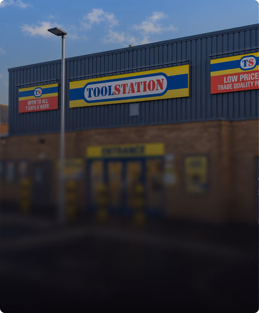Exterior of a Toolstation store with three signs on the upper building facade: the central sign says 'TOOLSTATION' with a blue, yellow, and red color scheme; the left sign reads 'OPEN TO ALL 7 DAYS A WEEK'; the right sign states 'LOW PRICES TRADE QUALITY PRODUCTS'. A streetlamp stands near the building against a partly cloudy sky.