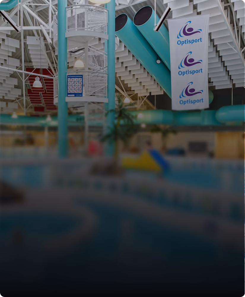 Indoor water park with teal water slides and a spiral staircase, featuring banners with the Optisport logo hanging from the ceiling.