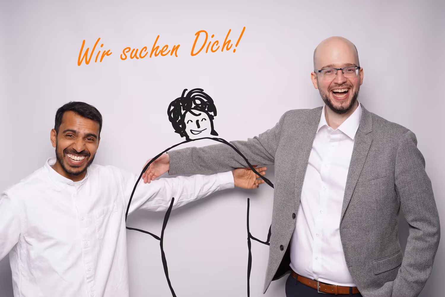 Zwei Männer lächeln in die Kamera und umarmen eine neutrale Comicfigur mit dem Schriftzug: „Wir suchen dich!“