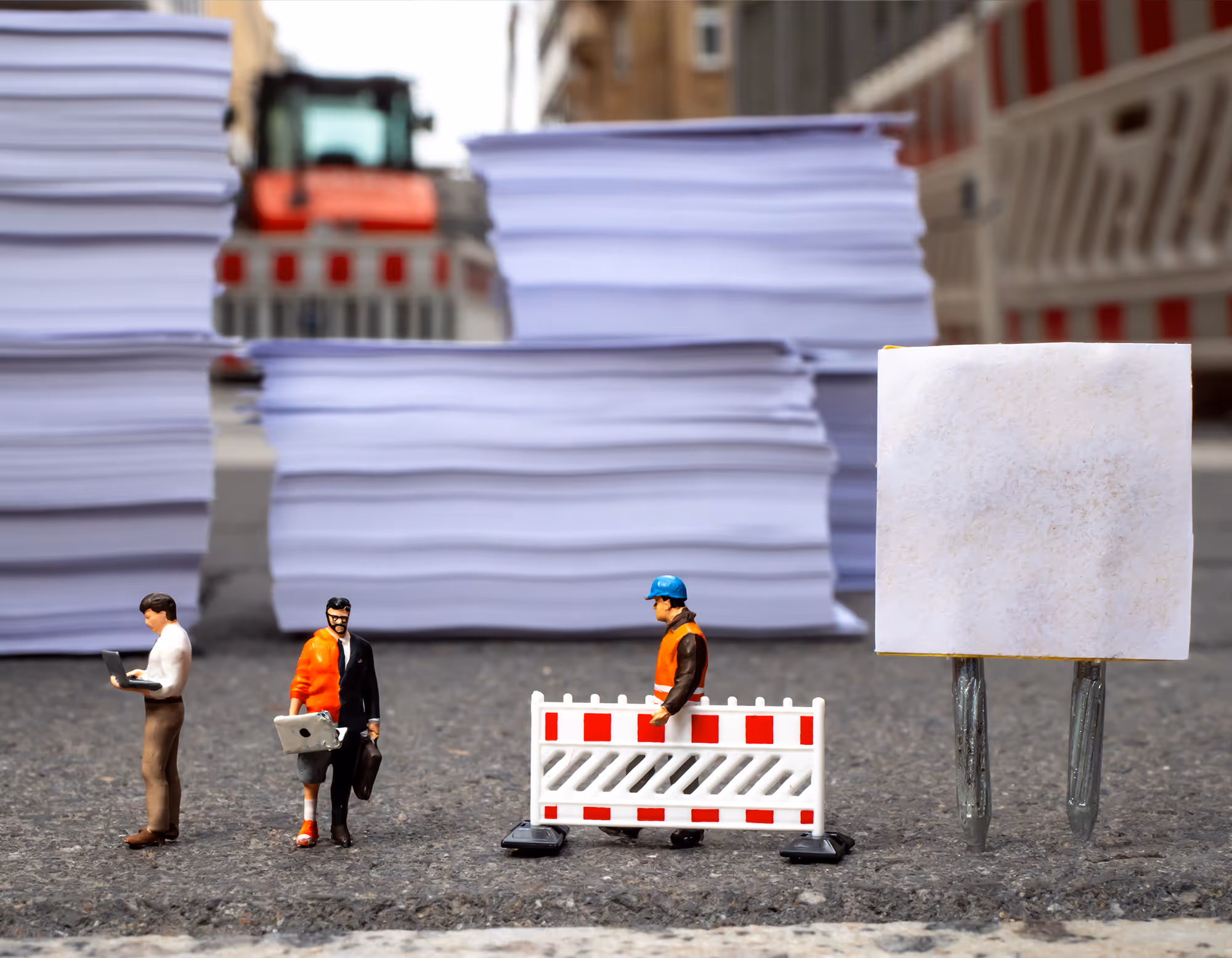 Miniaturfiguren von Bauarbeitern und Geschäftsleuten vor großen Papierstapeln und einer Baustellenabsperrung auf Asphalt.
