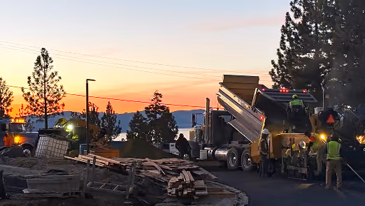 Construction crew paving a road at the Peak resort at dusk, with a dump truck and lumber in the foreground and a lake and mountains silhouetted at sunset.