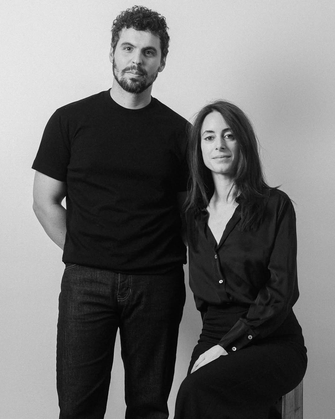Portrait en noir et blanc d’un homme debout et d’une femme assise, tous deux vêtus de noir, devant un fond clair. Ils sont les fondateurs et photographes de Sight by Sight.