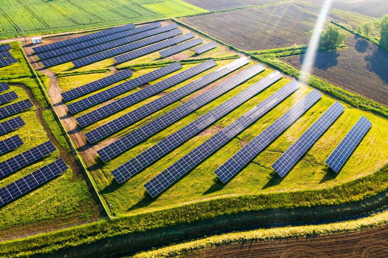 Aerial drone view of rows of solar panels installed across a green field, with dirt access tracks between rows and farmland in the background.