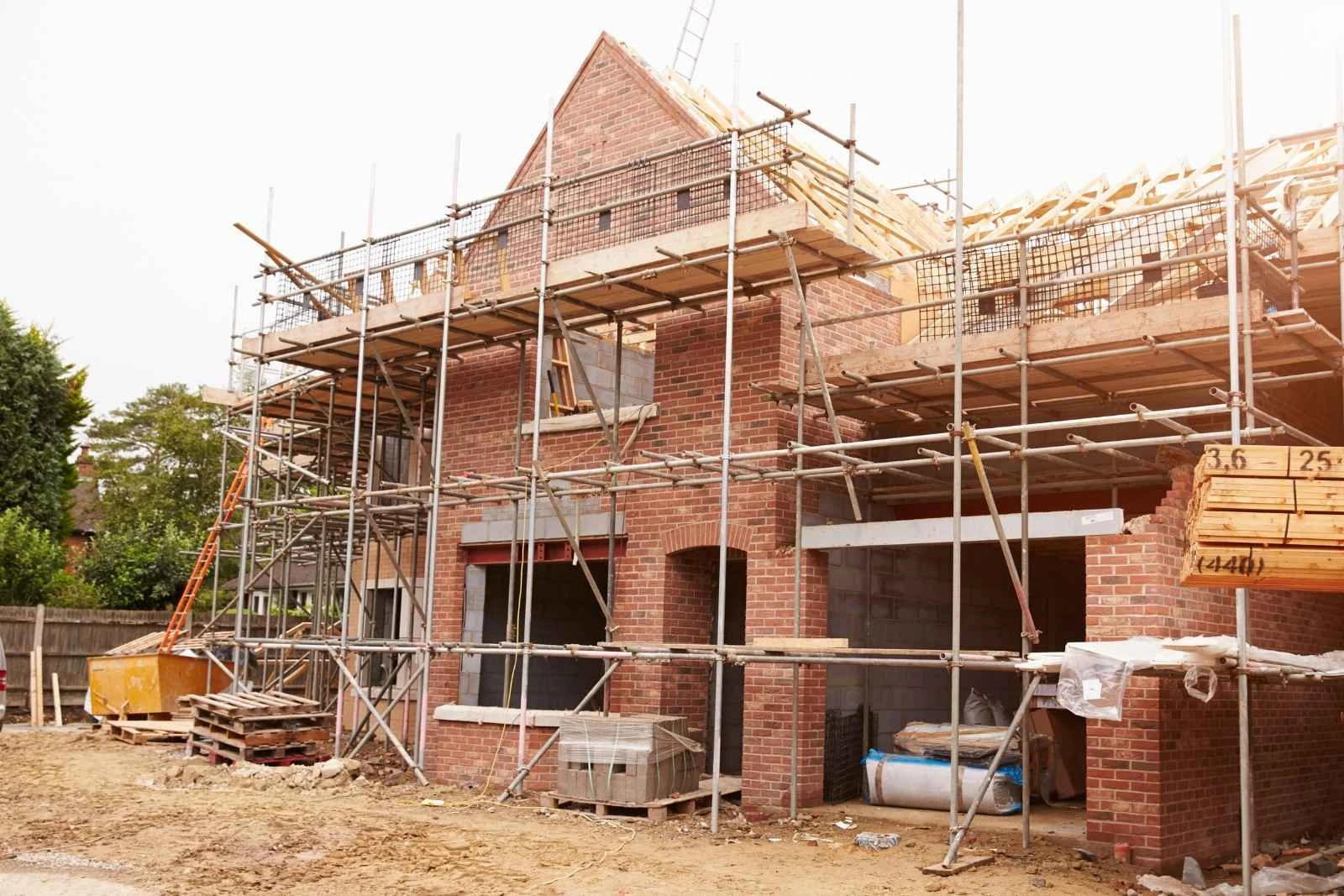 Small UK residential development site under construction — a part-built brick house wrapped in scaffolding, with exposed roof trusses, building materials, and a skip in the front garden