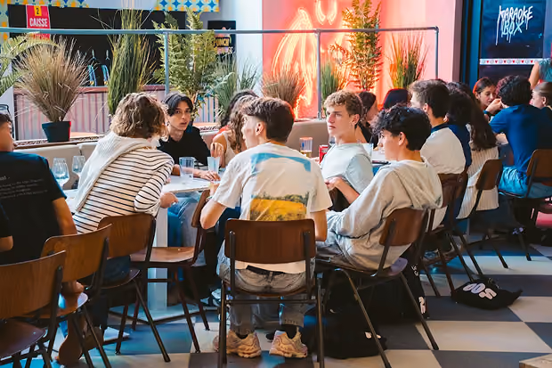 Jeunes assis autour de tables dans un café décoré avec des plantes et un néon rouge.