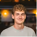 Jeune homme aux cheveux bouclés et t-shirt gris souriant devant un fond flou lumineux.