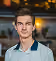 Jeune homme souriant avec une chemise à col bleu clair, arrière-plan flou avec lumière jaune.