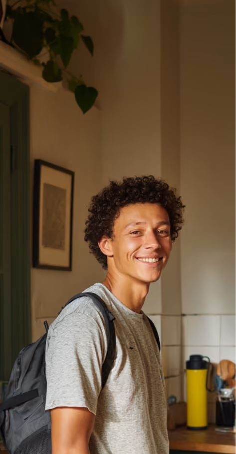Jeune homme souriant portant un t-shirt gris et un sac à dos dans une cuisine avec une bouteille jaune.