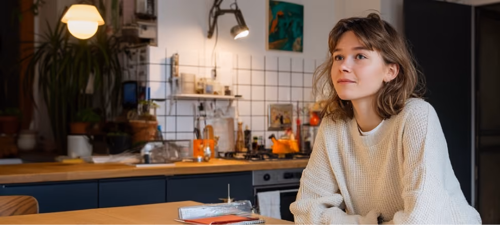 Jeune femme souriante en pull beige assise à une table en bois dans une cuisine moderne.