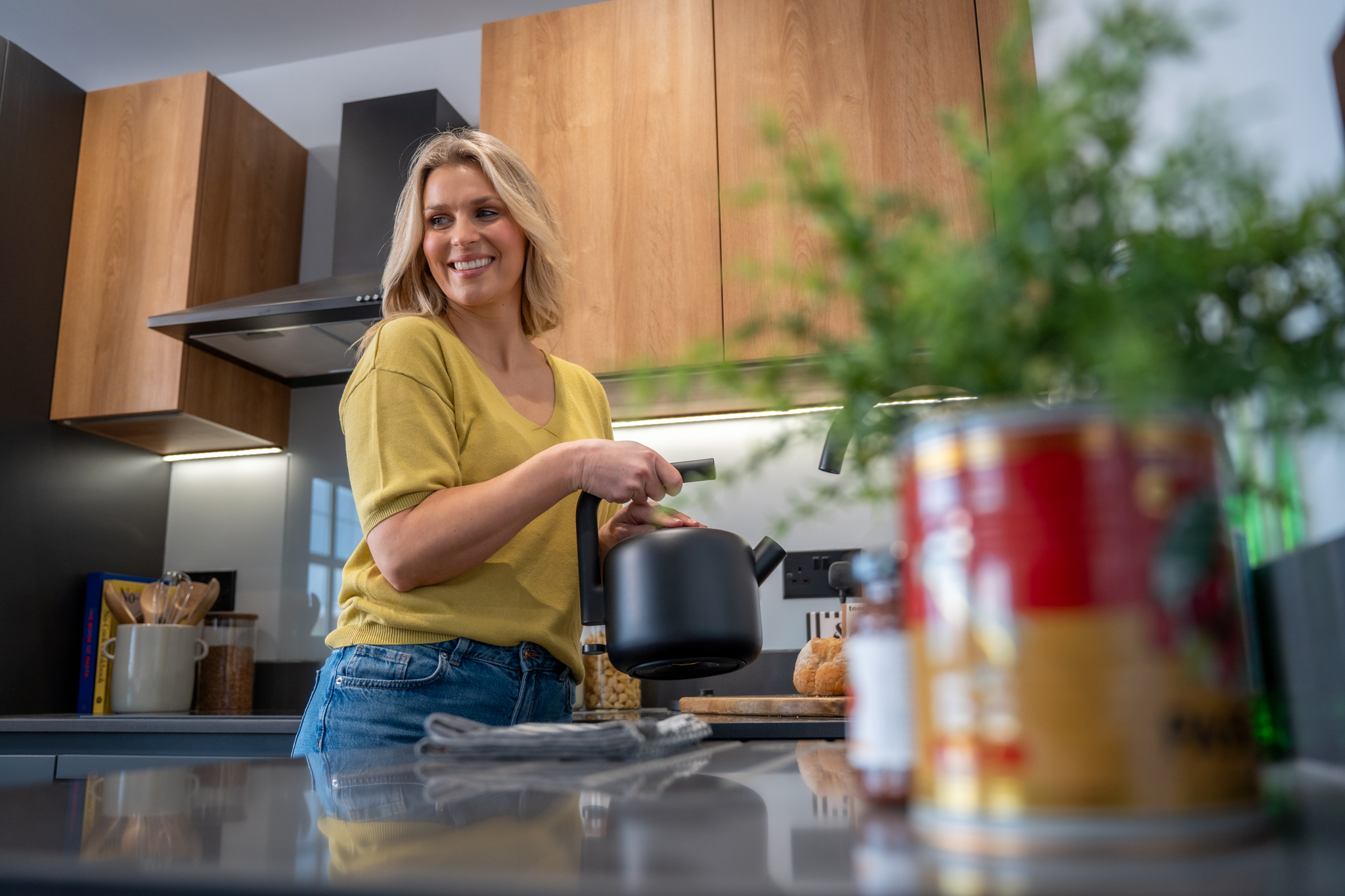 A woman smiling while holding a black kettle in a modern kitchen with wooden cabinets and blurred foreground plants and tins.