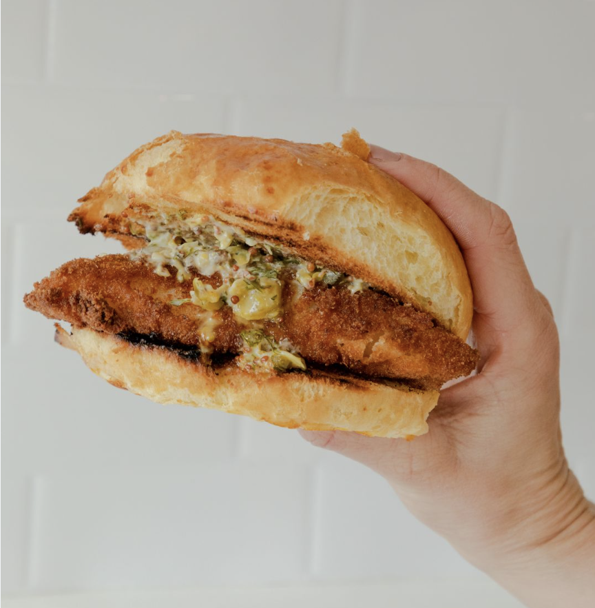 A hand holding a large fried fish sandwich with tartar sauce.