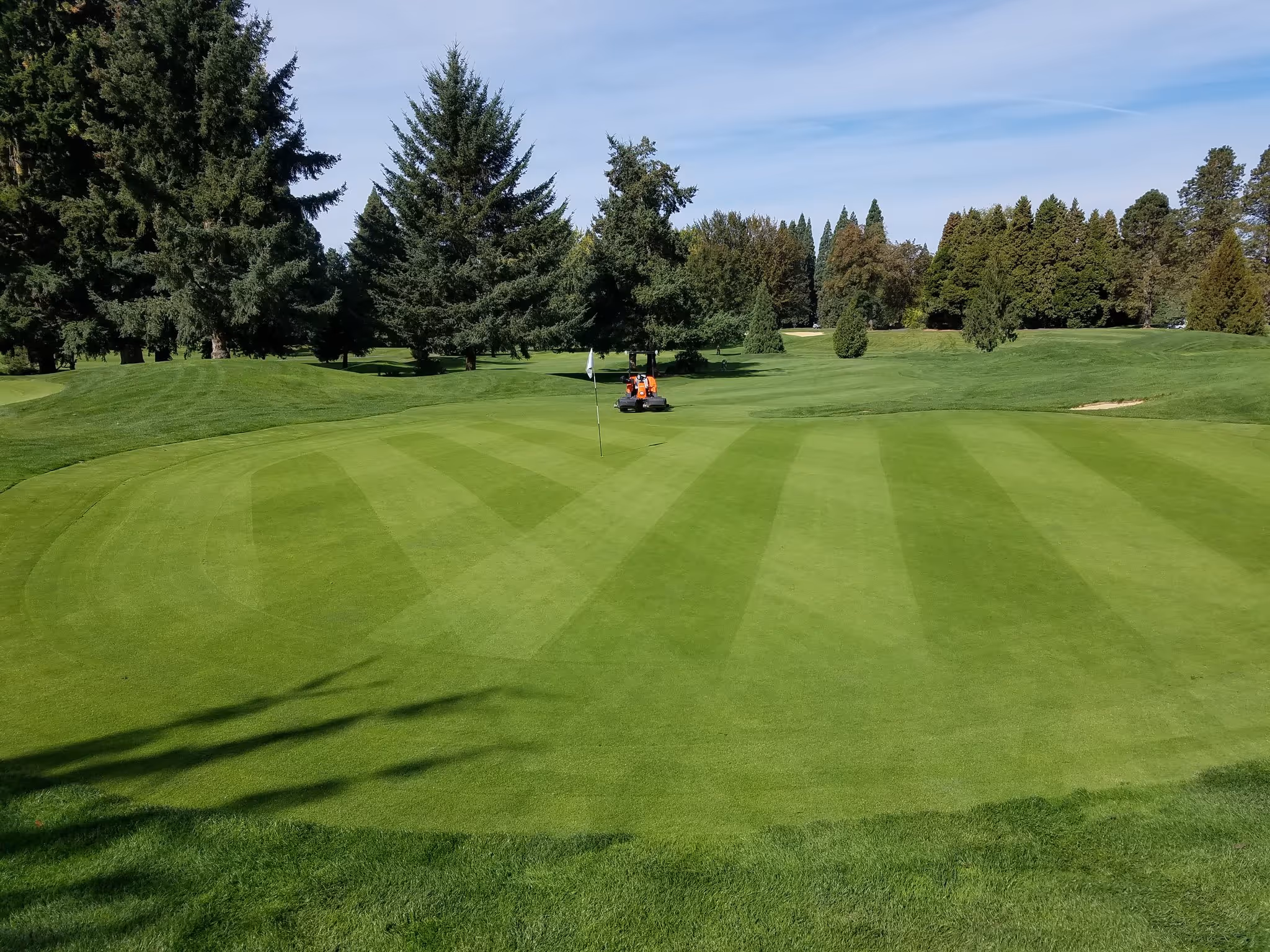 Columbia Edgewater golf course green with striped mowing patterns, flagstick, and a landscaping vehicle near tall pine trees under a blue sky.