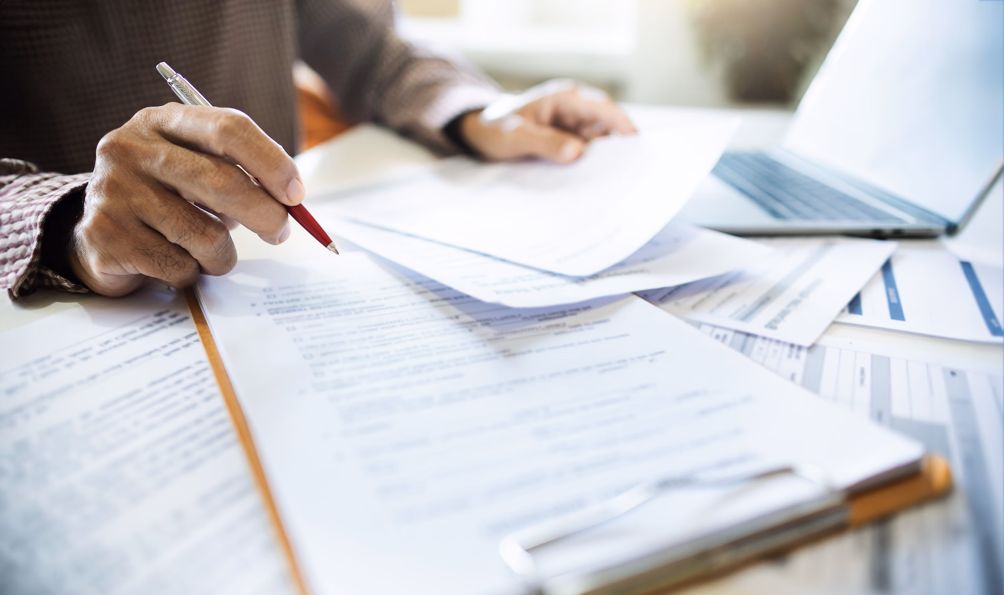 Person holding a red pen reviewing and organizing multiple printed documents on a desk with a laptop nearby.