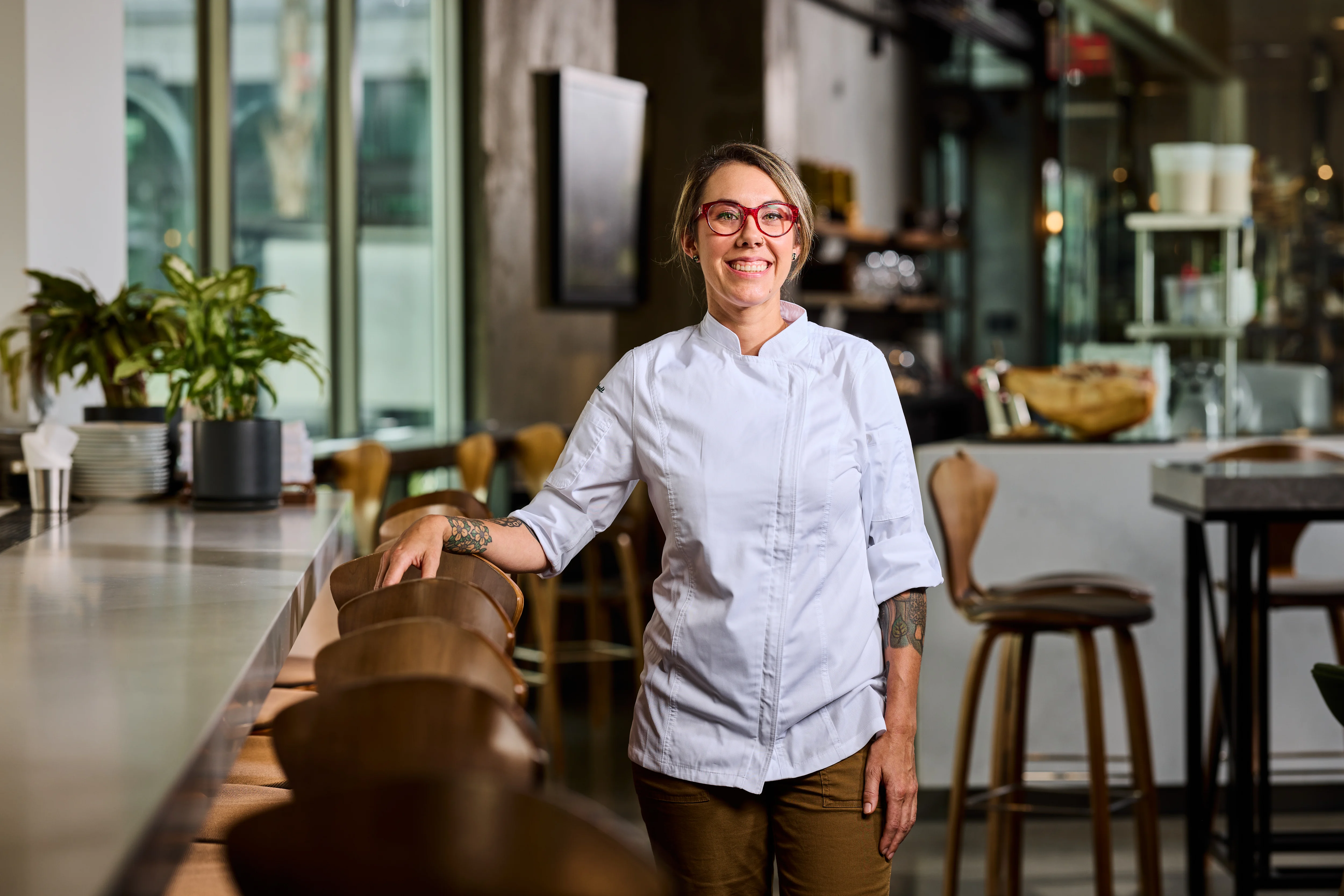 Smiling female chef with tattoos and red glasses standing in a modern restaurant interior.