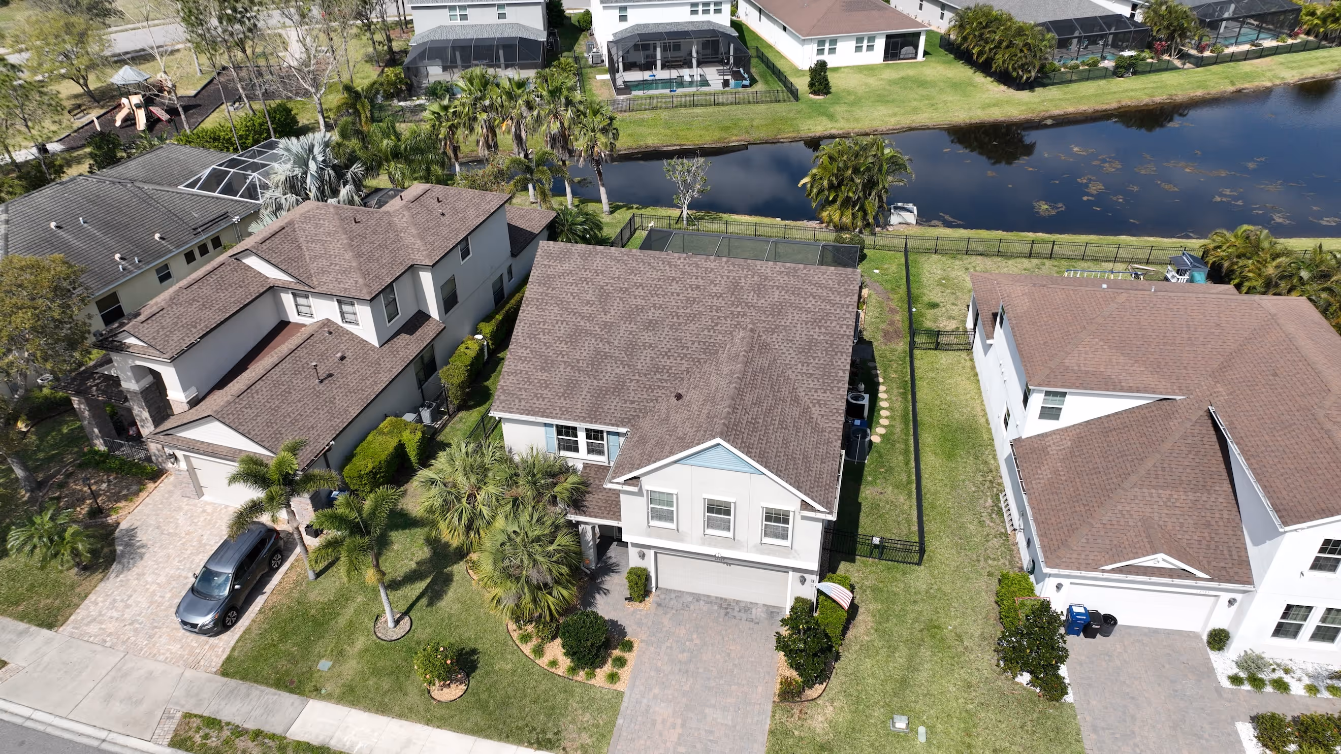 Aerial view of suburban houses with brown roofs, green lawns, palm trees, and a water canal behind the properties.