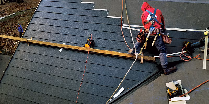Worker in red hoodie and safety harness installing dark roof tiles on a sloped roof with tools and safety ropes.