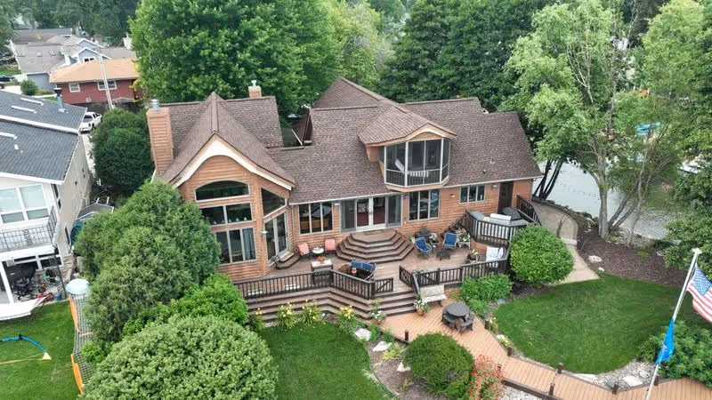 Aerial view of a large wooden house with a spacious deck, surrounded by green trees and lawn next to a water body.