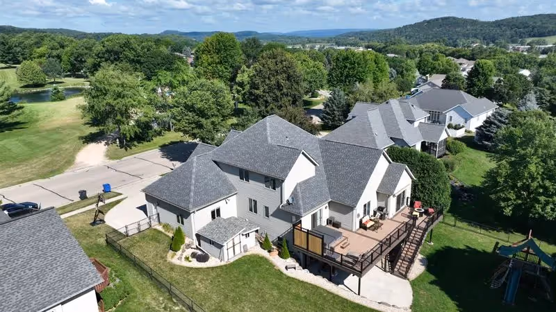 Aerial view of a large suburban house with gray roofing, a wooden deck with chairs, surrounded by a green lawn and other houses and trees in the background.