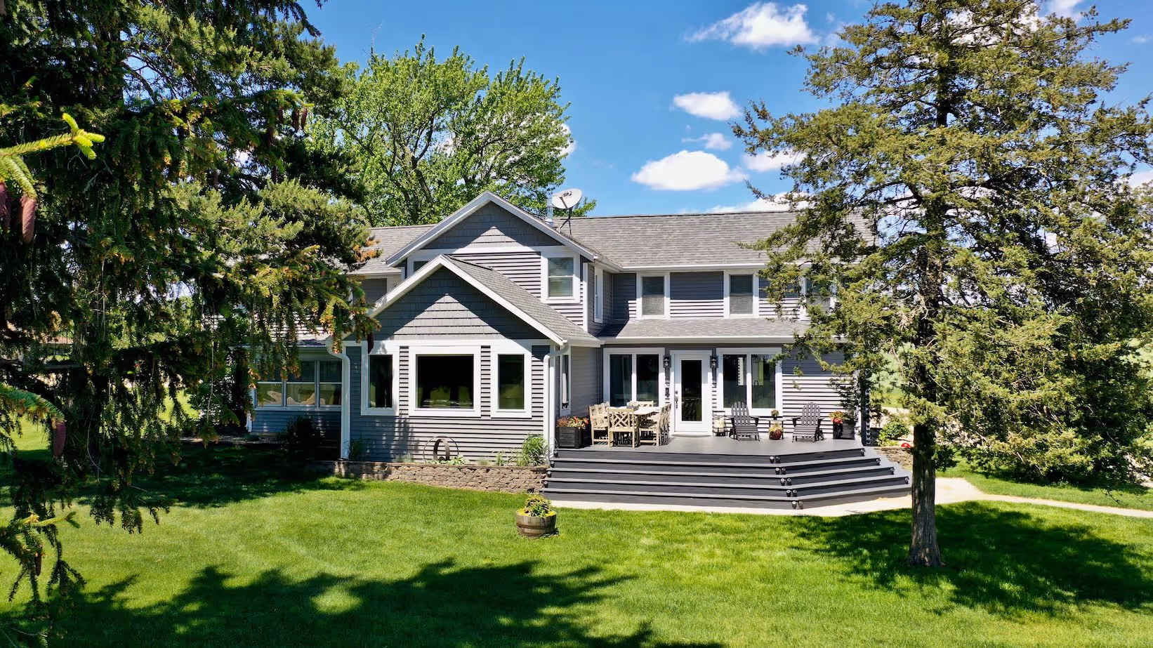 Two-story gray house with white trim, large porch with outdoor furniture, surrounded by green lawn and trees under a blue sky.