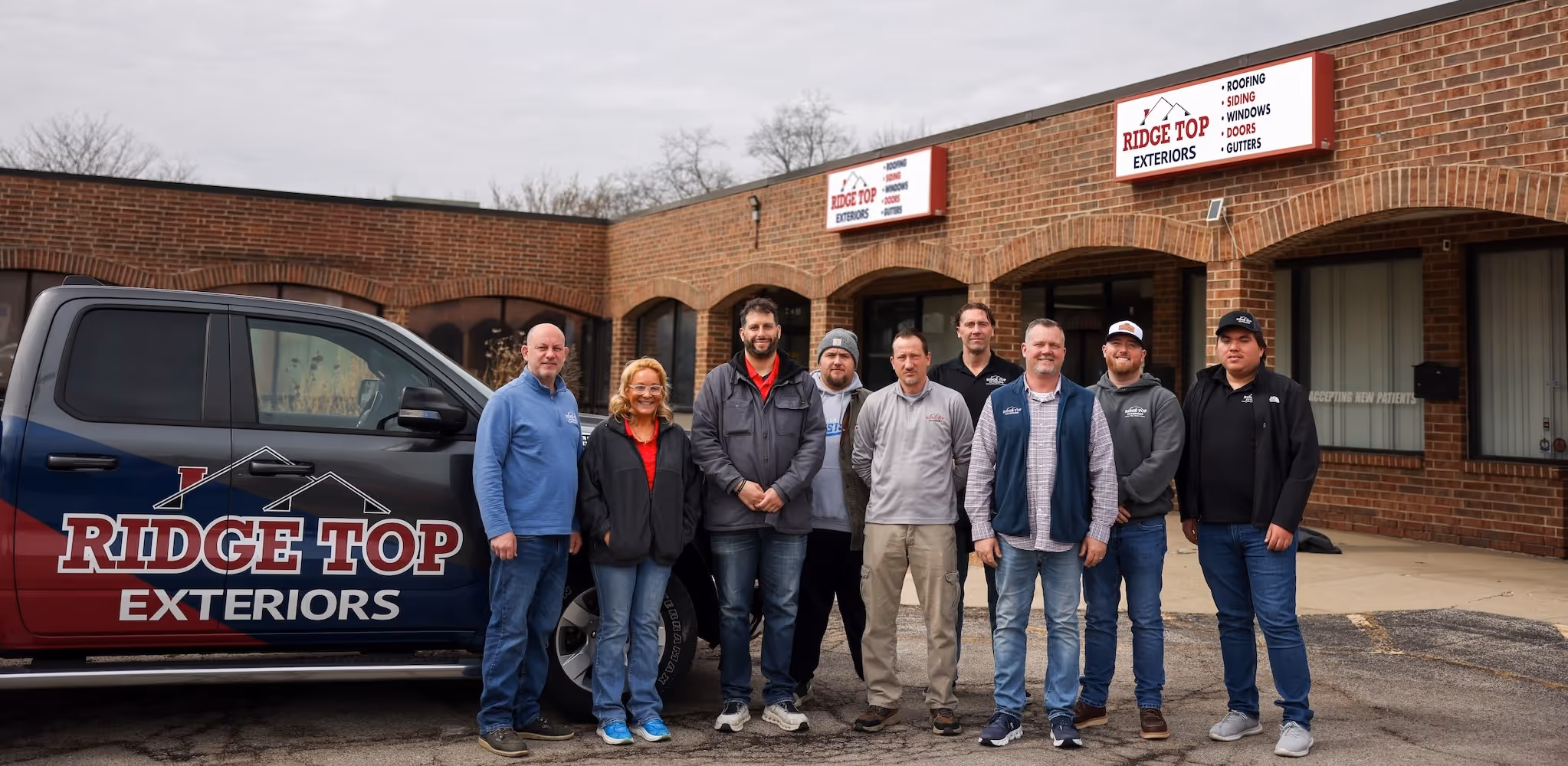 Six men wearing Ridge Top Exteriors polo shirts standing in front of a brick building with a glass door and window.