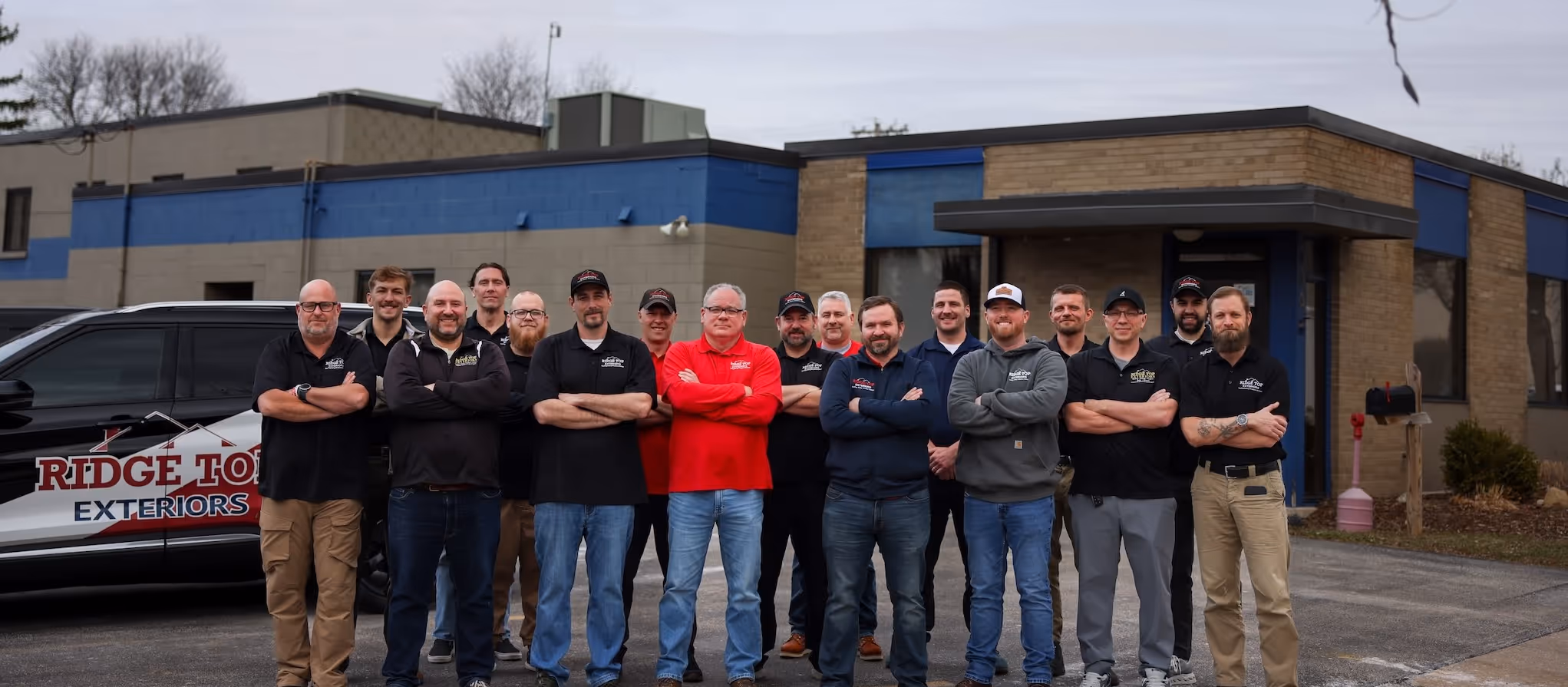 Group of fifteen men standing outdoors in front of a building and white truck, wearing red, black, and blue company shirts.