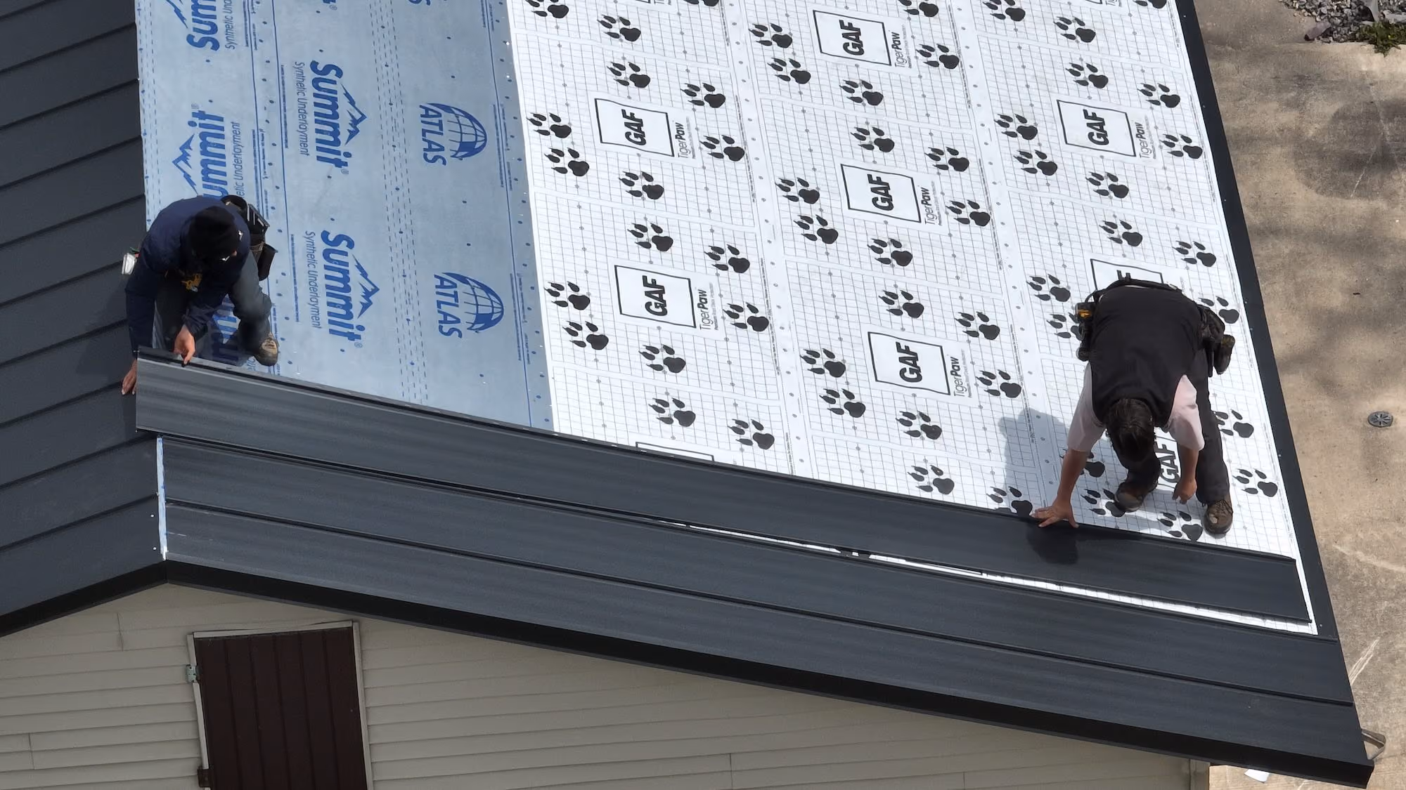 Two workers installing dark metal roofing panels over synthetic underlayment on a house roof.
