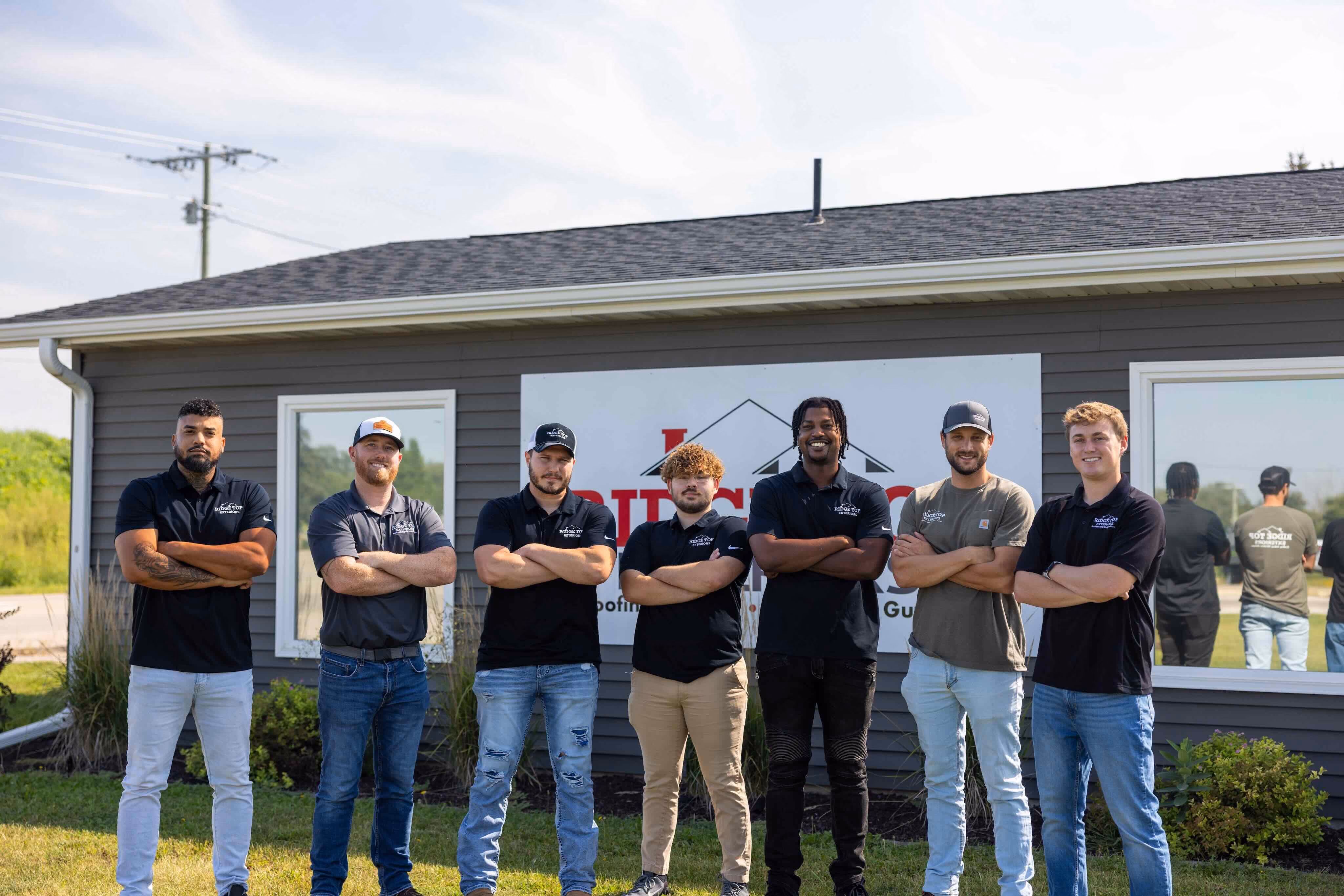 Seven men standing outside in front of a gray building with a sign behind them, posing with arms crossed and smiling.