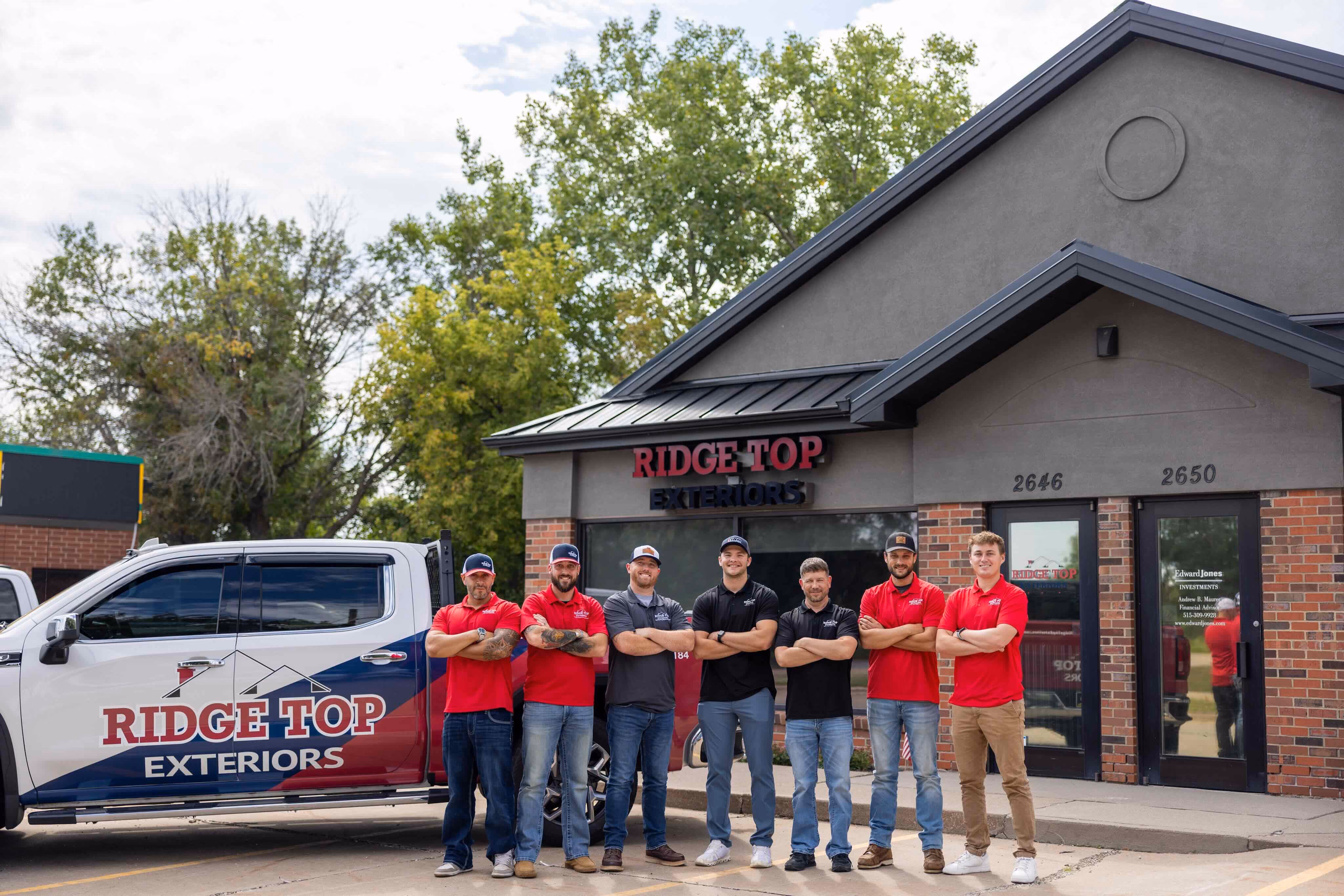 Seven men wearing red and black Ridge Top Exteriors shirts standing with arms crossed in front of a Ridge Top Exteriors building and pickup truck under a partly cloudy sky.