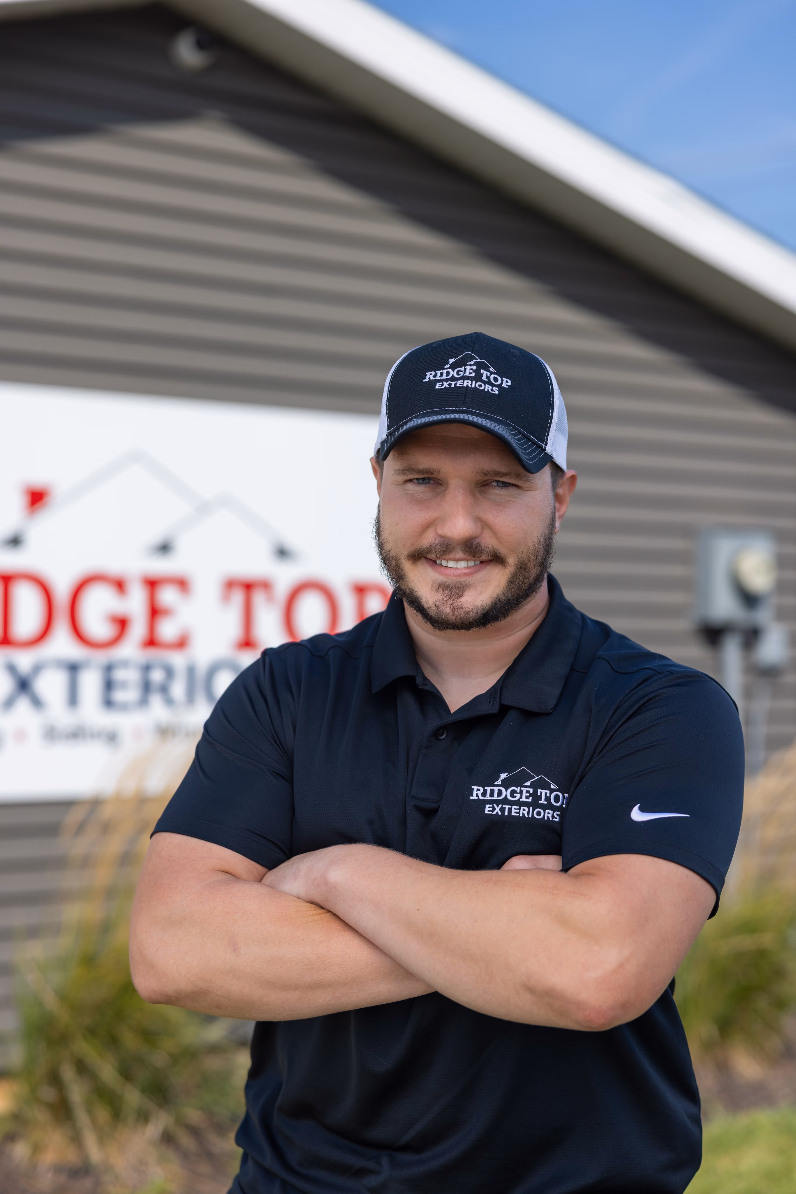 Man with a beard and mustache wearing a black Ridge Top Exteriors cap and matching polo shirt, standing with arms crossed in front of a building with a Ridge Top Exteriors sign.