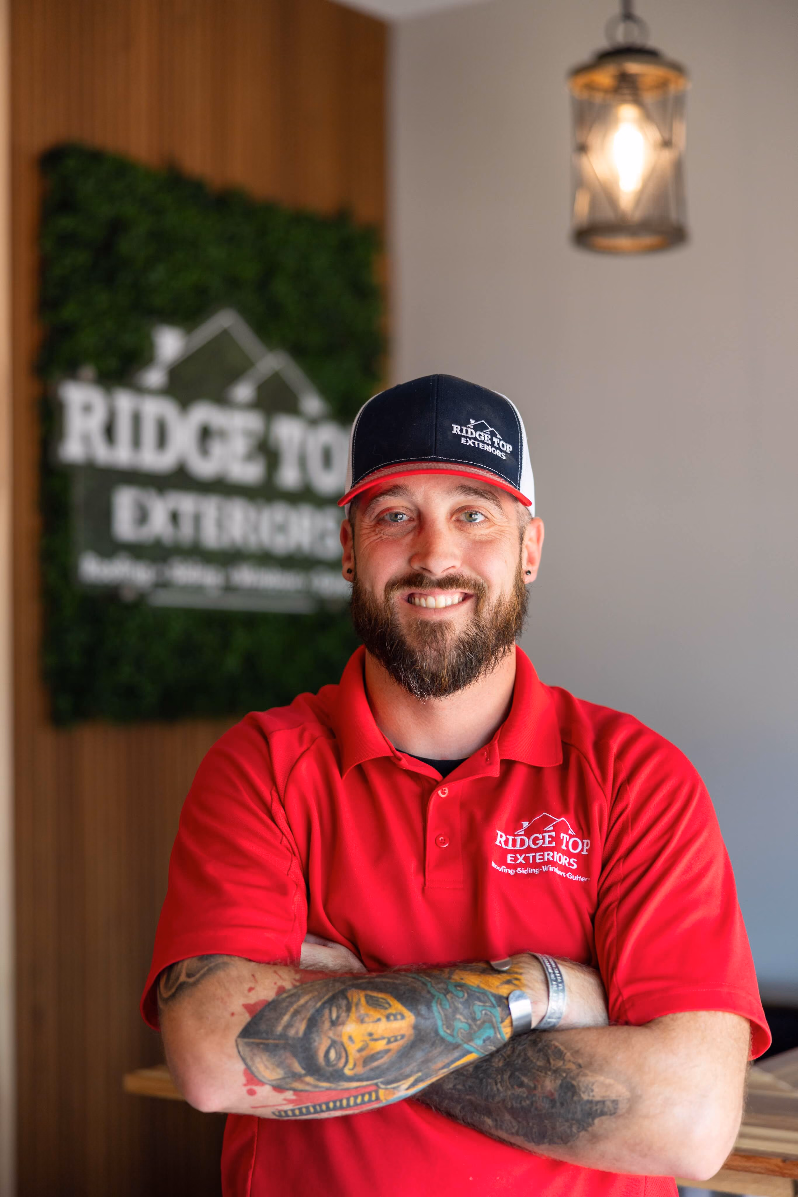 Randy Scadden - Bearded man with a tattooed forearm wearing a red Ridge Top Exteriors shirt and matching cap, standing with arms crossed and smiling indoors in front of a green and wood Ridge Top Exteriors sign.