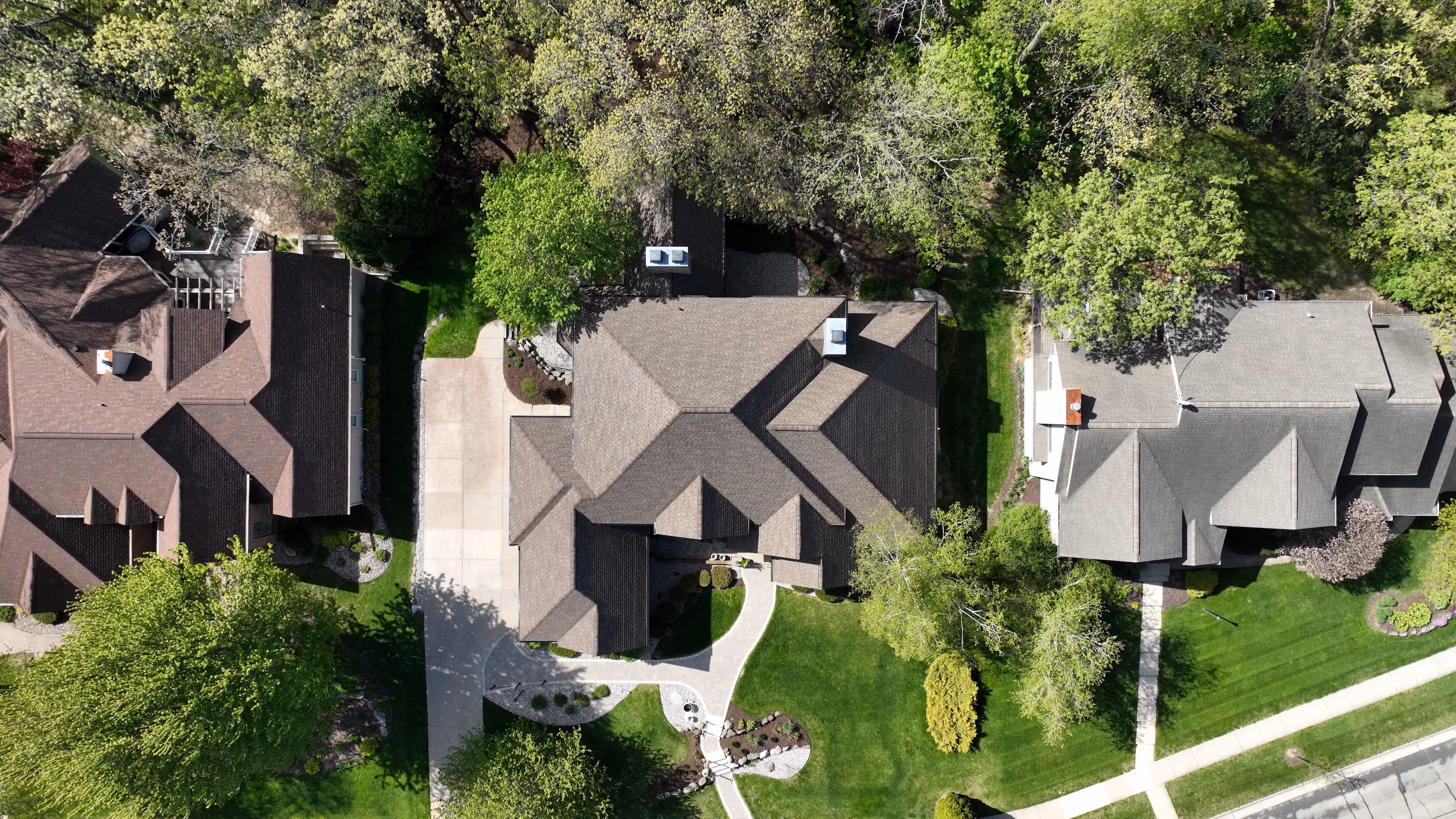 Aerial view of suburban homes with brown and gray architectural asphalt shingle roofs surrounded by mature trees