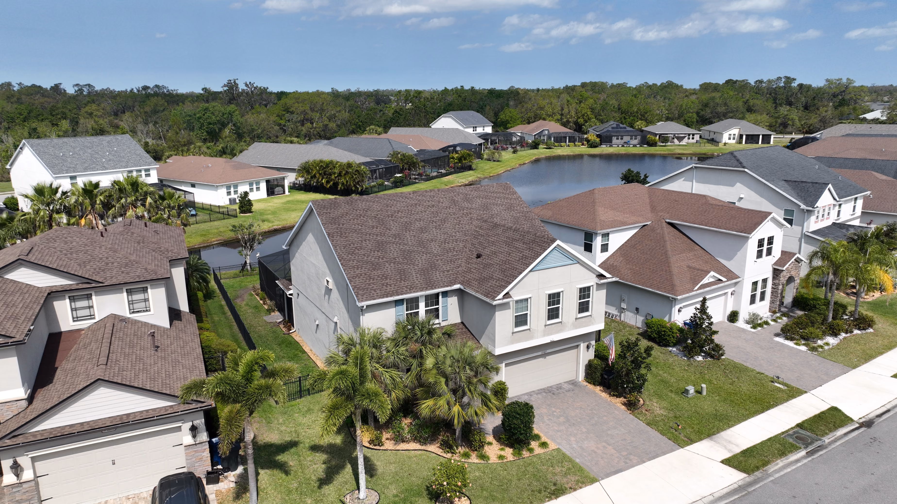 Aerial view of a two-story suburban home with a dark shingle roof, paver driveway, palm trees, and a community pond in the background.