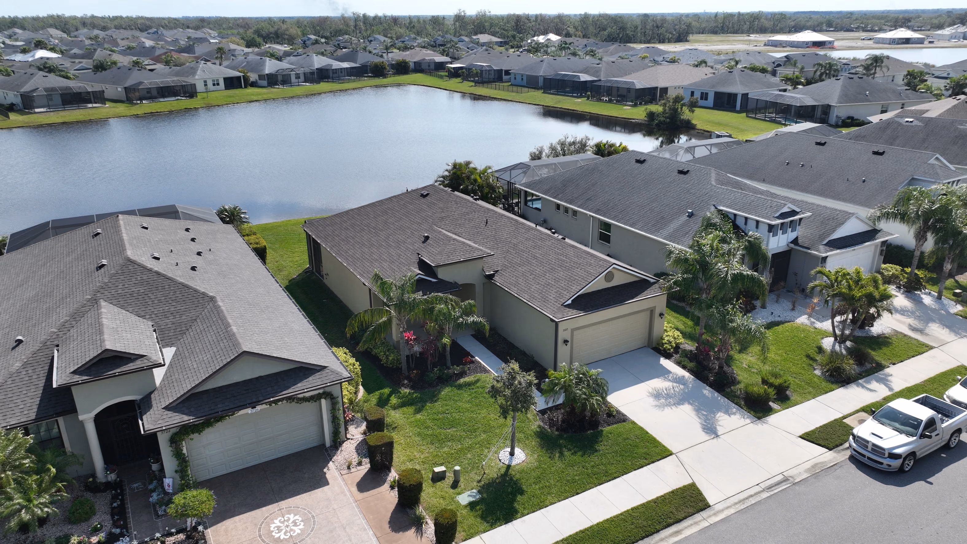 Overhead drone view of a single-story ranch home with an attached garage and manicured lawn backing onto a neighborhood retention lake.
