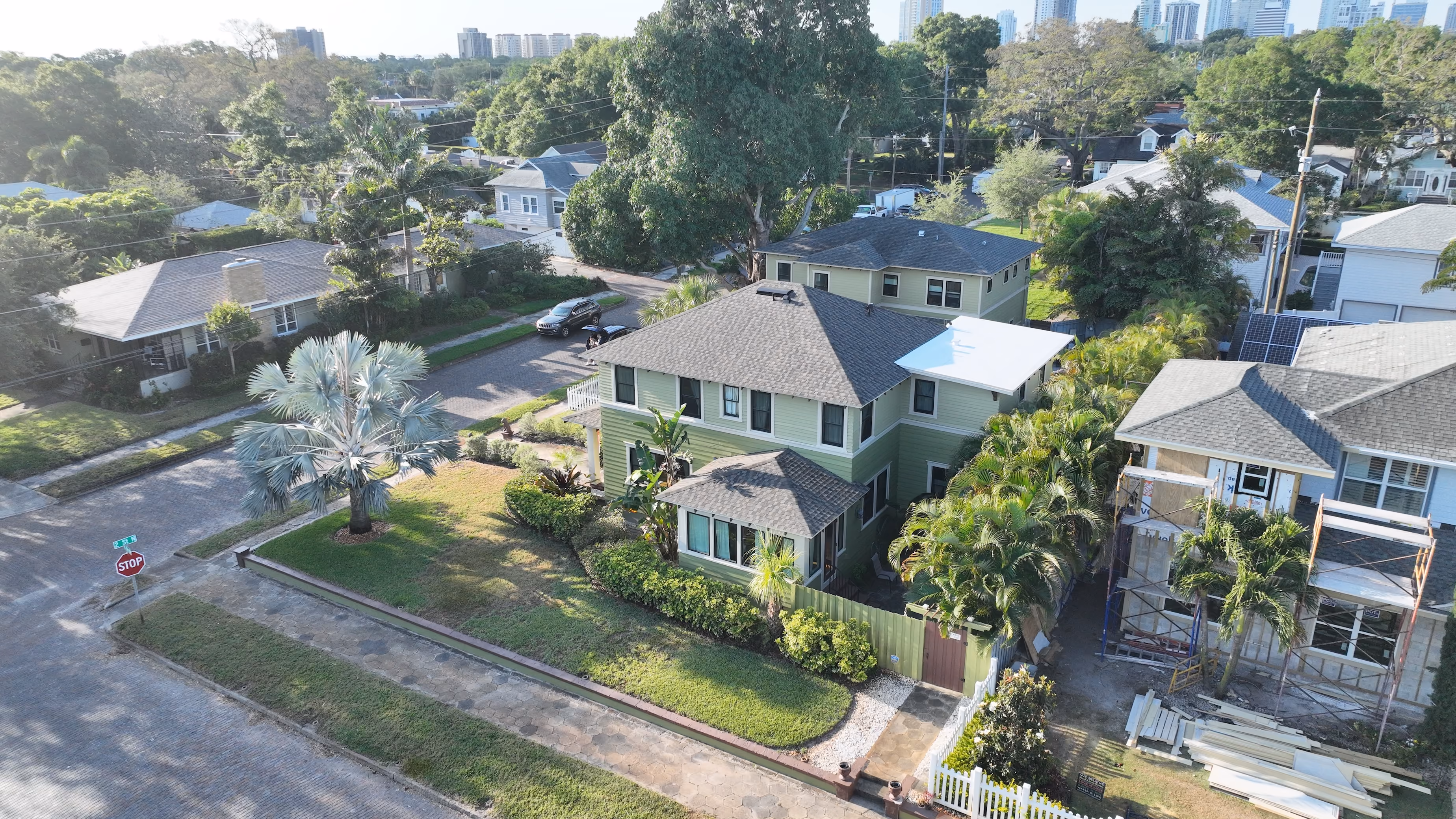 Aerial view of a two-story light green Craftsman-style house with a dark shingle roof, corner yard with a silver fan palm, and neighboring homes along a paved street.