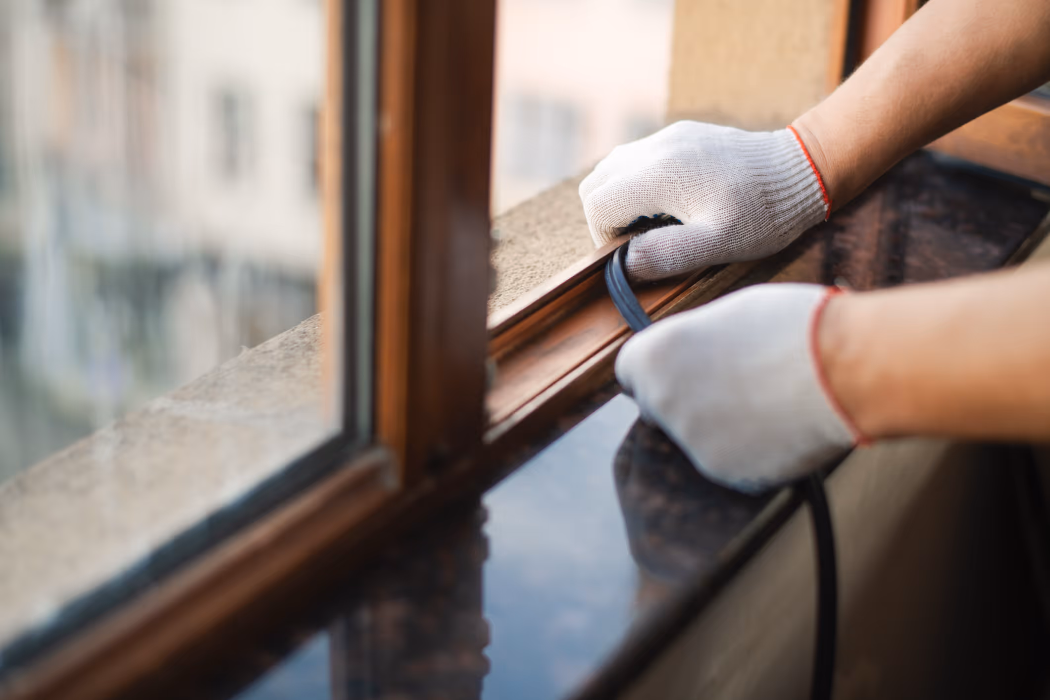 A gloved technician inspects and services a wooden window frame, checking seals and weatherstripping for air leaks that drive up monthly energy costs.
