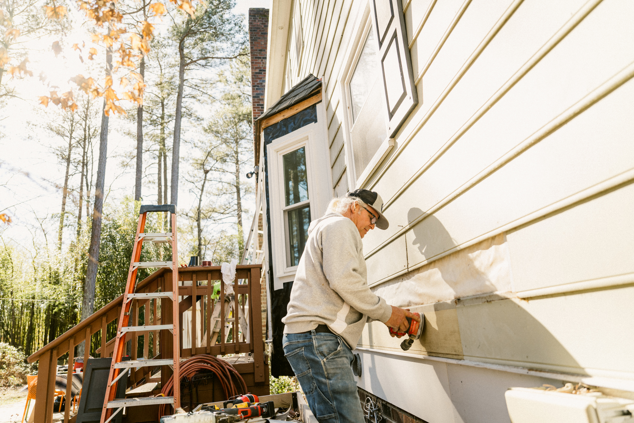 A contractor uses a power tool to repair vinyl siding panels on a residential home exterior, with ladders and equipment visible on the job site.