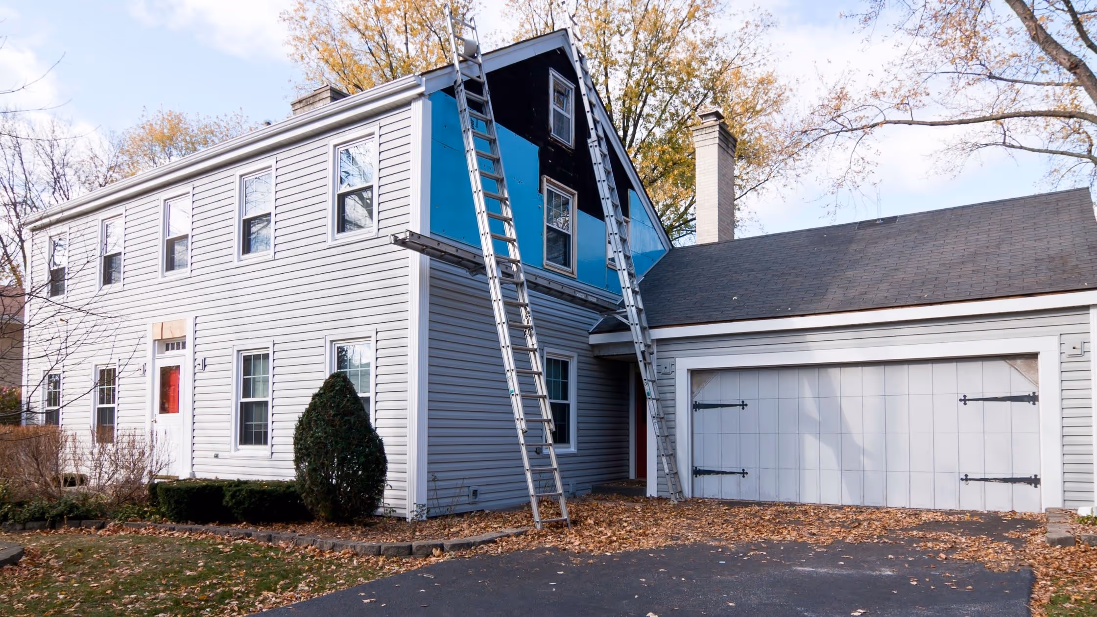 A two-story residential home mid-siding installation, with ladders leaning against the exterior and blue house wrap visible where old panels have been removed.