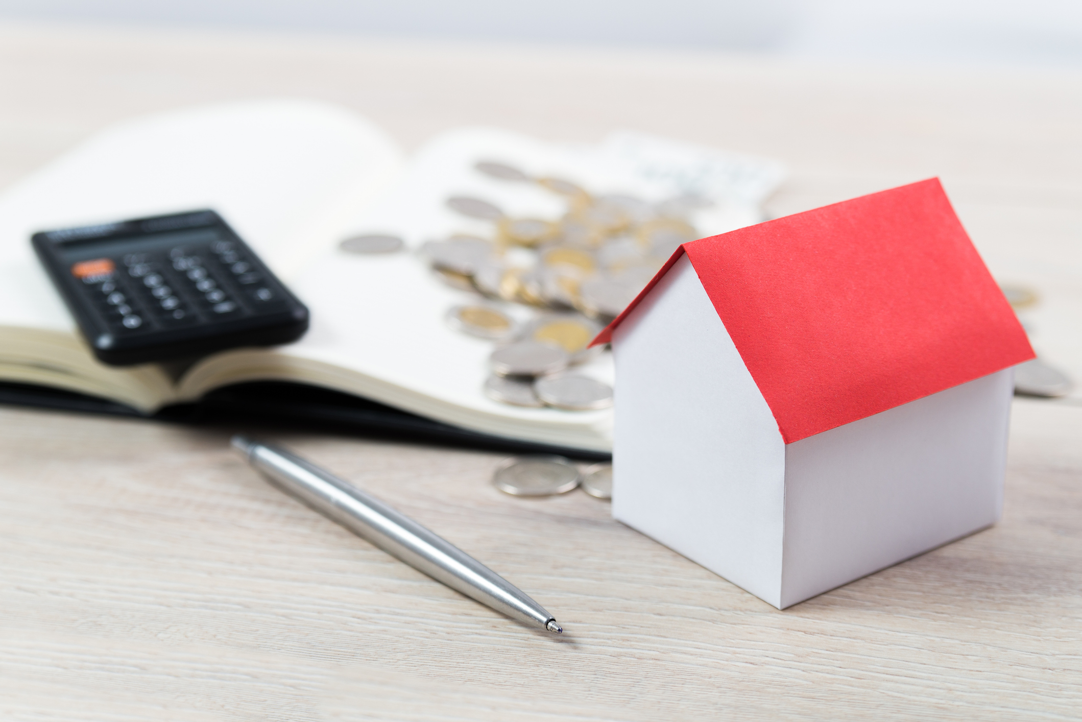 A small paper house model with a red roof next to a pen, calculator, and coins on an open notebook.