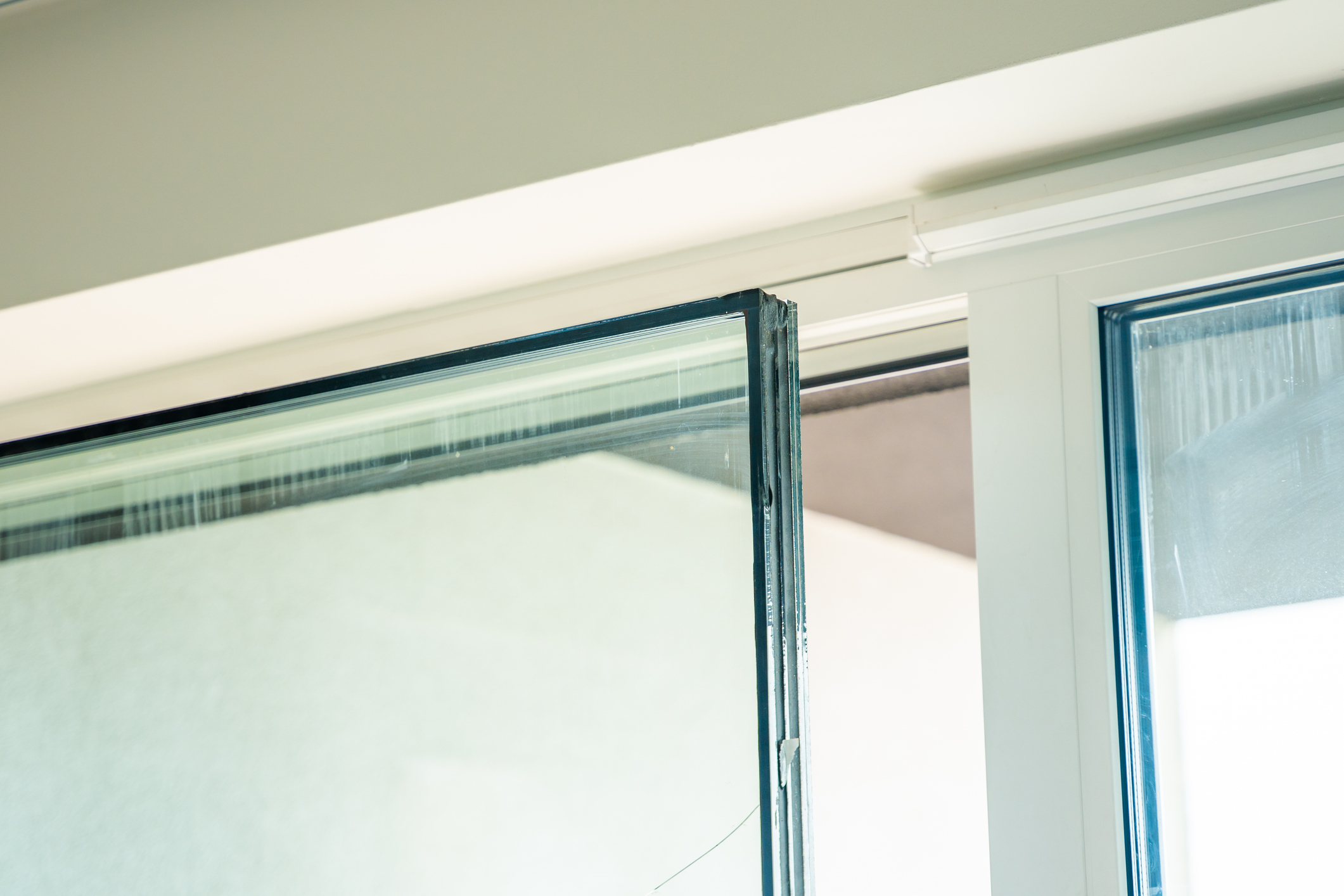 A close-up interior shot of a triple-pane window unit showing the three distinct glass layers and sealed spaces visible at the edge of the glass, with a white vinyl frame on the right and a light gray ceiling above.