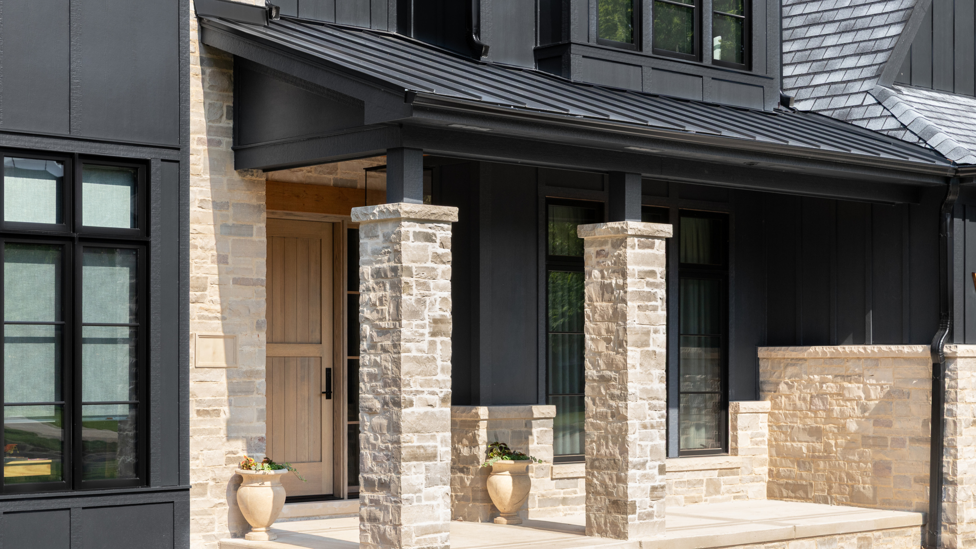 Modern home exterior with black board and batten siding, stone porch columns, a standing seam metal porch roof, and a wood front door.