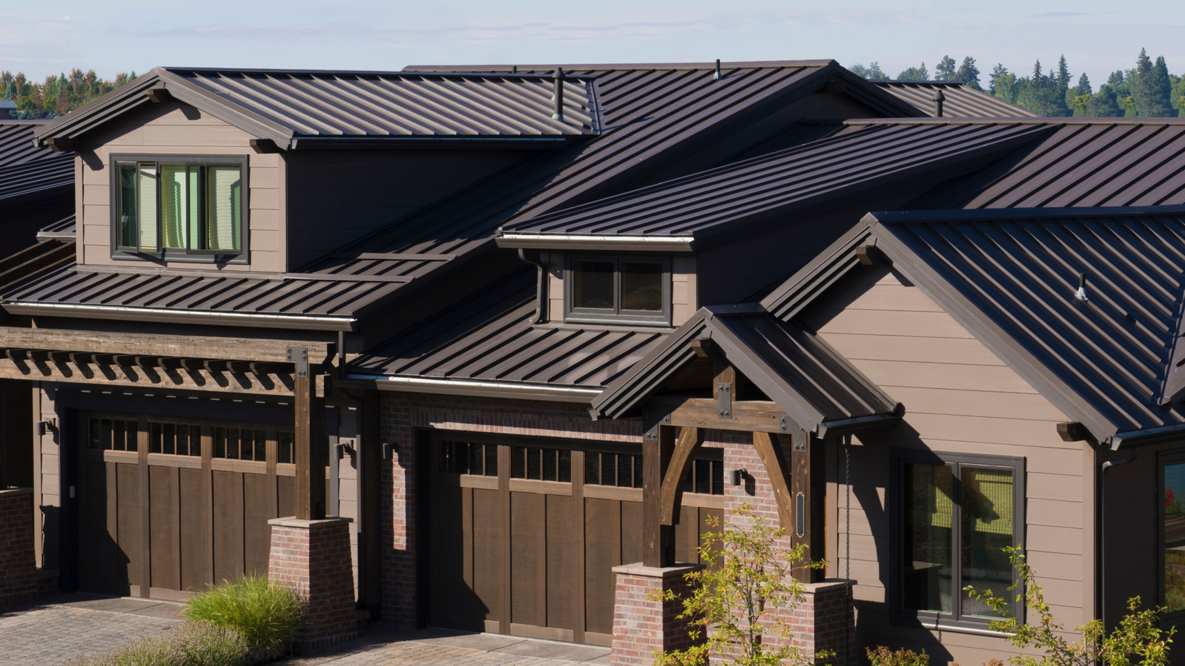 Craftsman-style home with a dark standing seam metal roof, multiple roof valleys, tan siding, and wood carriage-style garage doors.