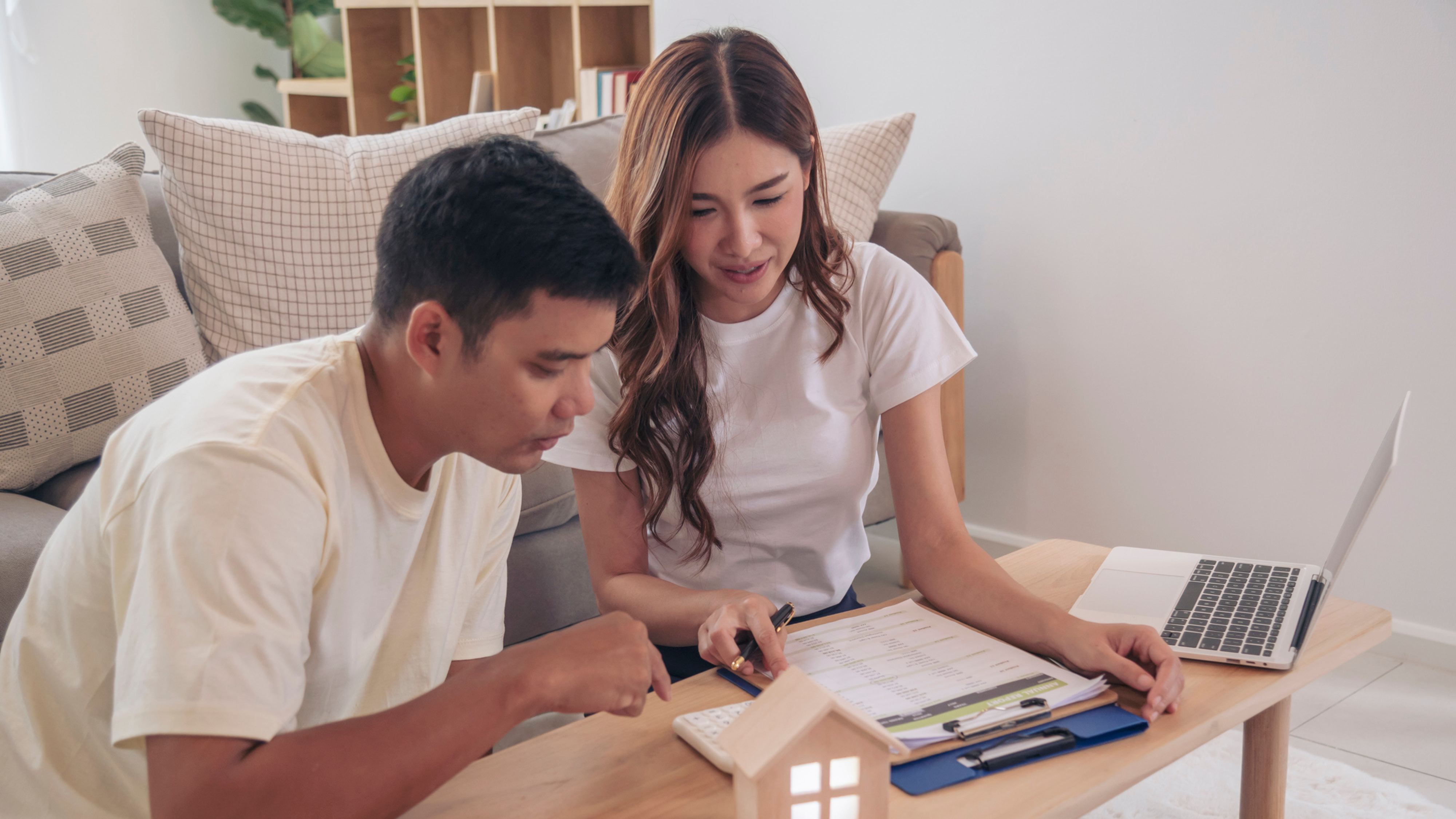 Young couple at a living room coffee table going over paperwork on a clipboard, with a calculator, laptop, and small wooden house model nearby.