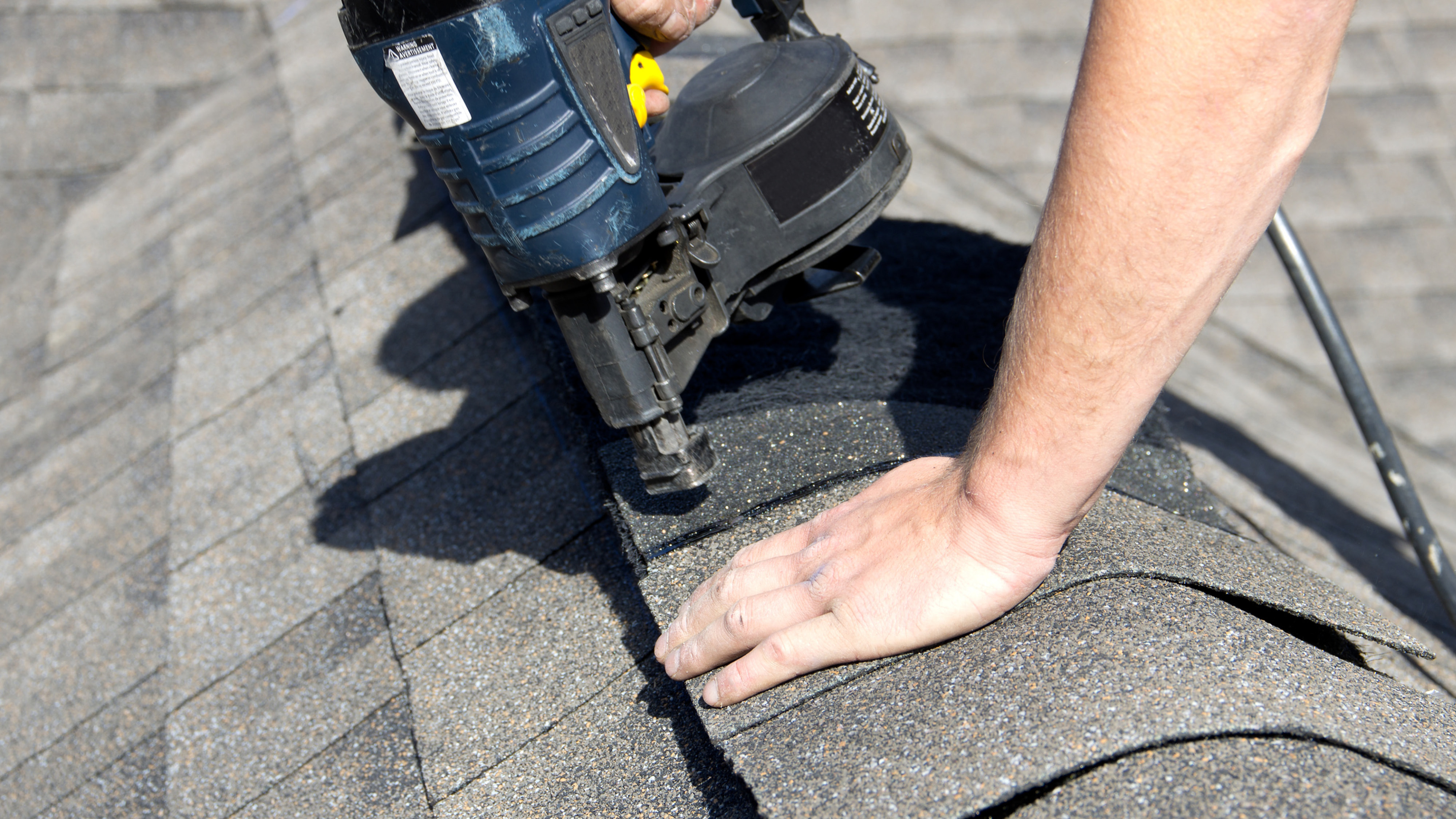 Close-up of a roofer's hand holding a gray asphalt shingle in place while fastening it to the roof ridge with a pneumatic nail gun.