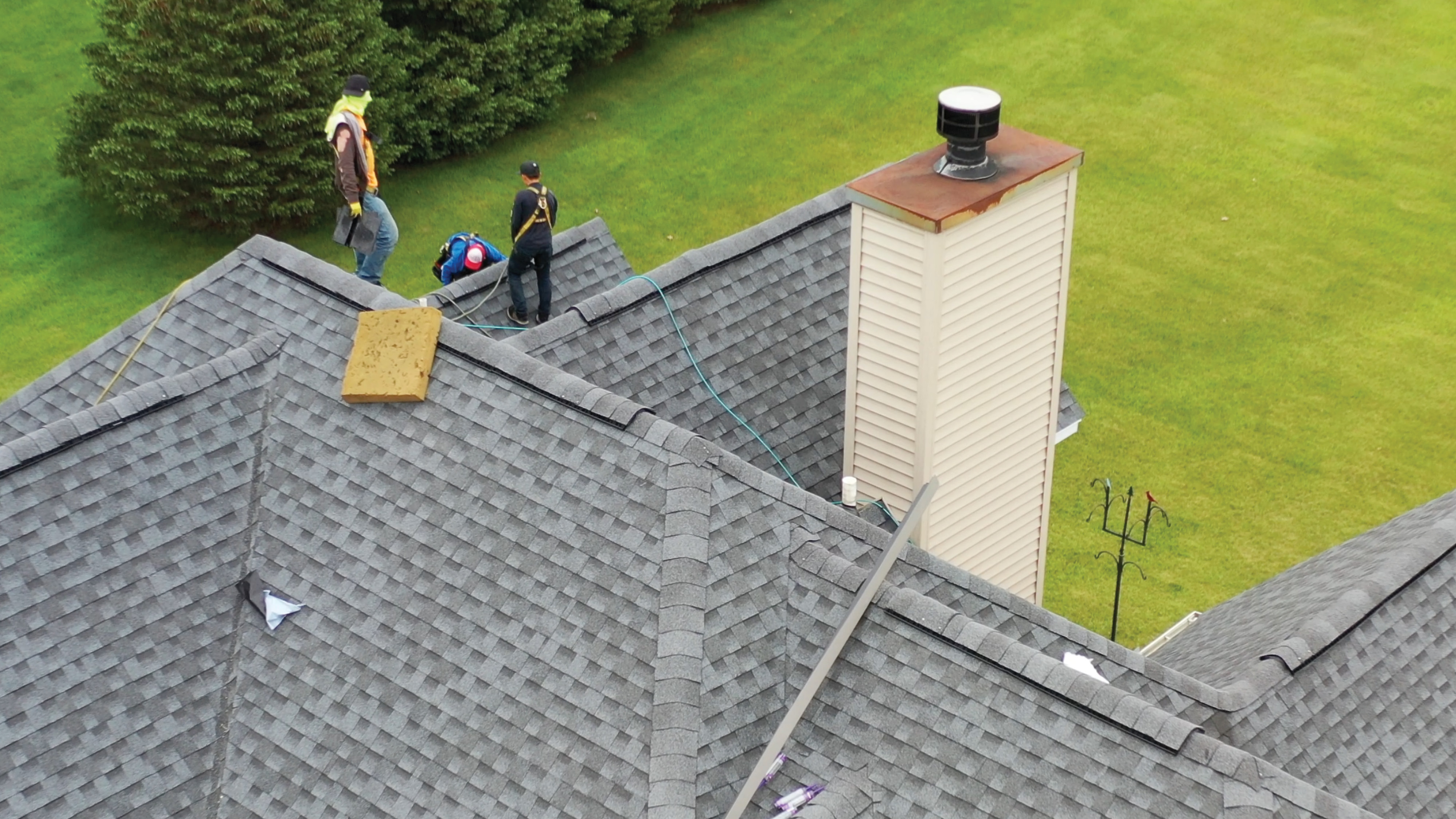 Aerial view of three roofers in safety harnesses working on a gray asphalt shingle roof with multiple ridges and a sided chimney.