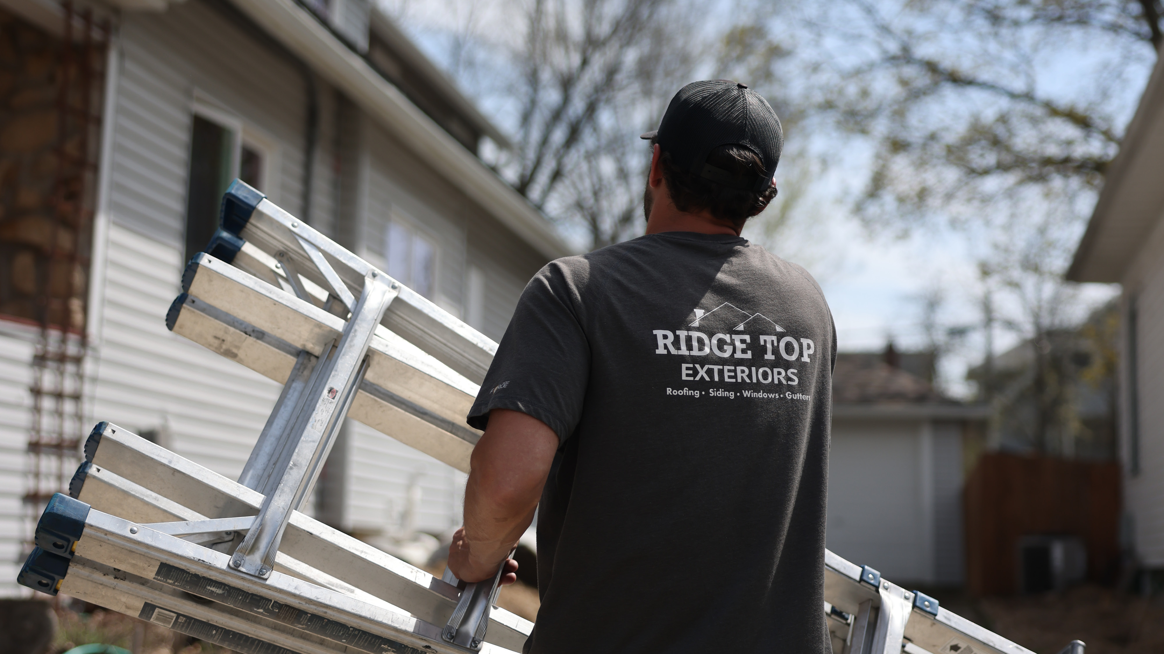 Ridge Top Exteriors crew member in a cap and branded t-shirt carrying a stack of aluminum ladders toward a home with white vinyl siding.