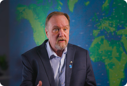Man in a navy blazer and light blue shirt speaking, with a world map blurred in the background.