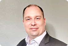 Smiling man in a dark suit jacket and light shirt against a plain gray background.