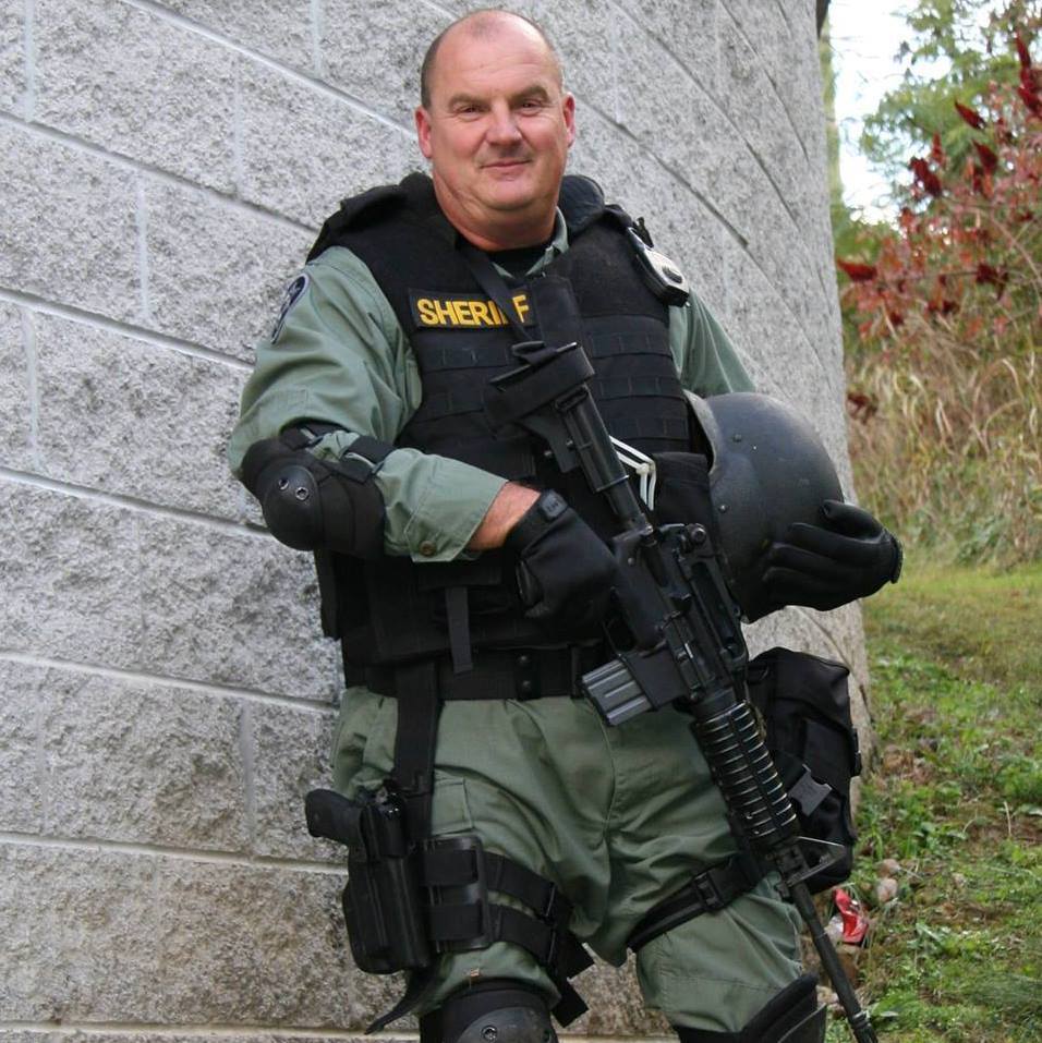 Sheriff in tactical gear holding a helmet and rifle, standing against a stone wall outdoors.