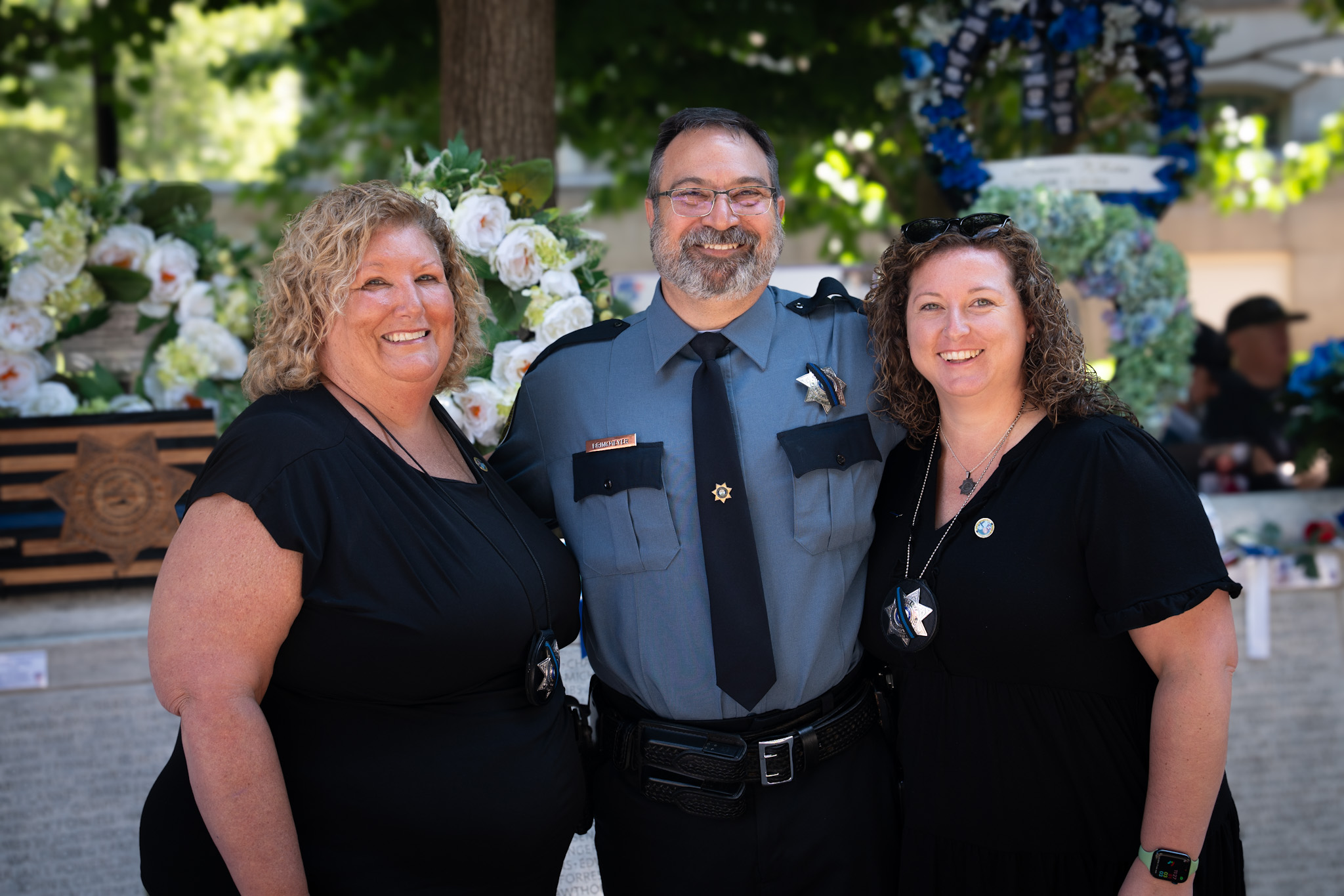 Three smiling individuals, two women and one man in police uniform, standing outdoors near floral memorials.