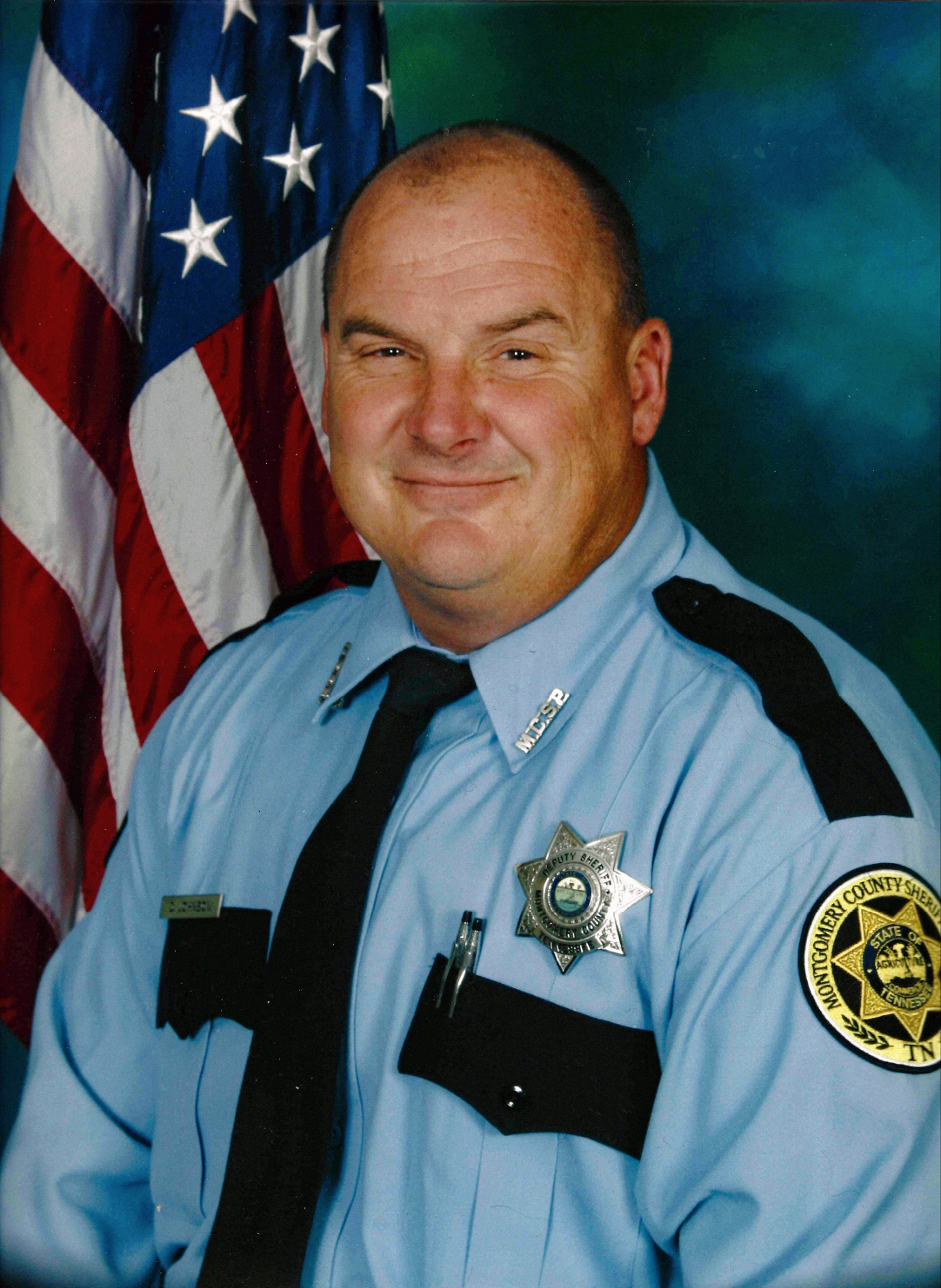 Smiling male deputy sheriff in blue uniform with badges and American flag background.